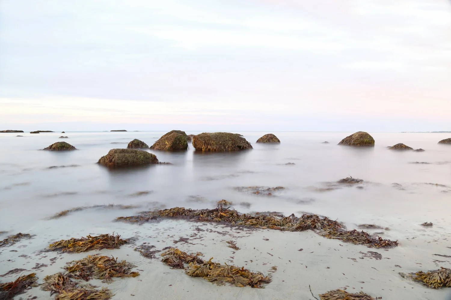 A long exposure photo of a calm beach with rocks in the water and seaweed on the sandy shore, under a cloudy sky.