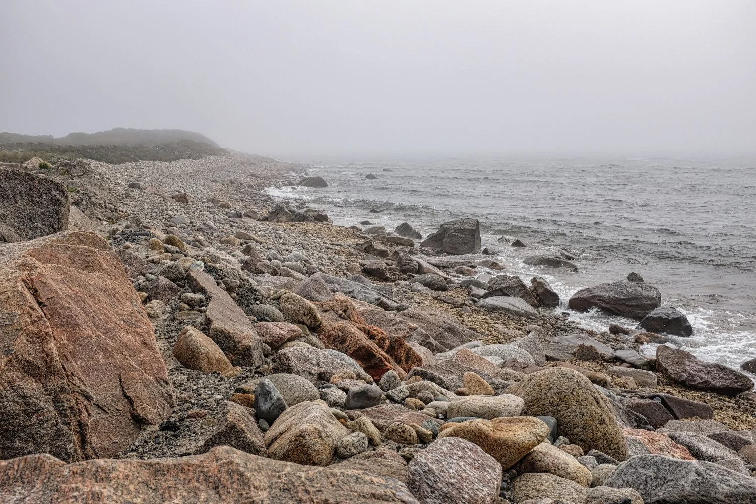 A rocky beach with large stones along the shoreline, foggy weather, overcast sky, and calm ocean waves.
