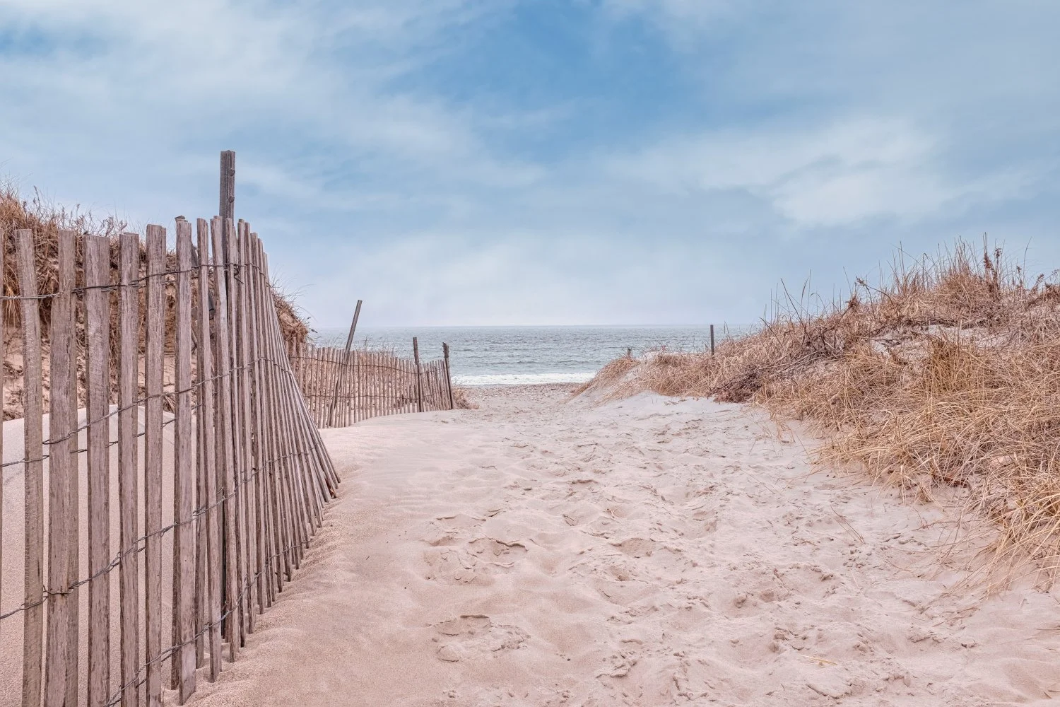 Beach scene with sandy pathway, wooden fences, sand dunes with dry grass, ocean in the background, partly cloudy sky