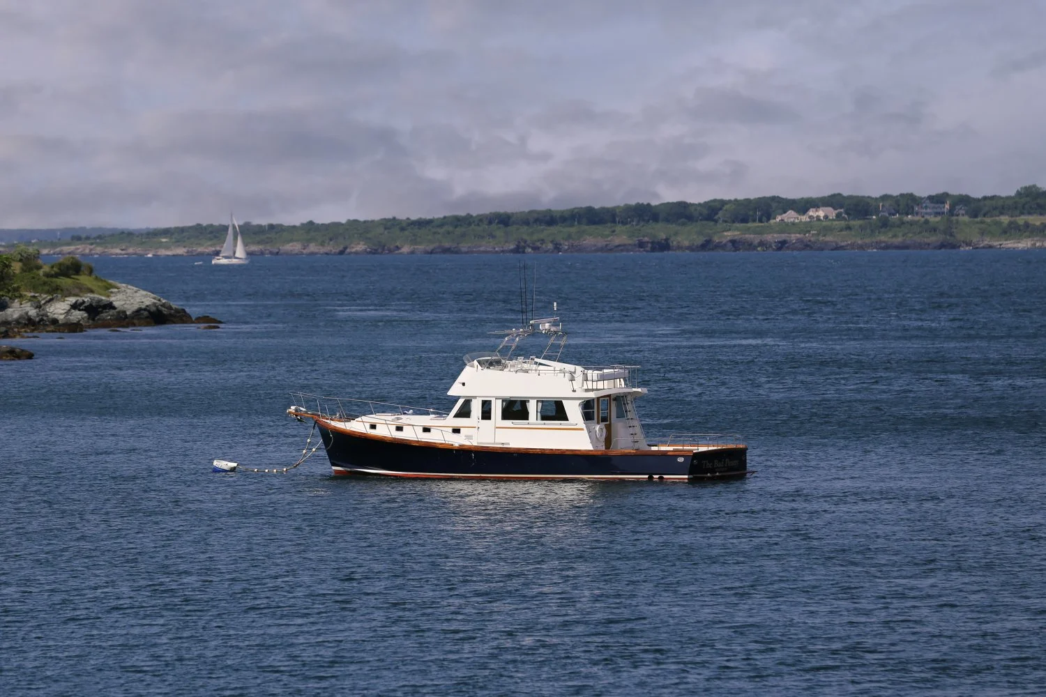 A white and black motorboat floating on calm blue waters near a rocky shoreline with a sailboat in the distance under a cloudy sky.