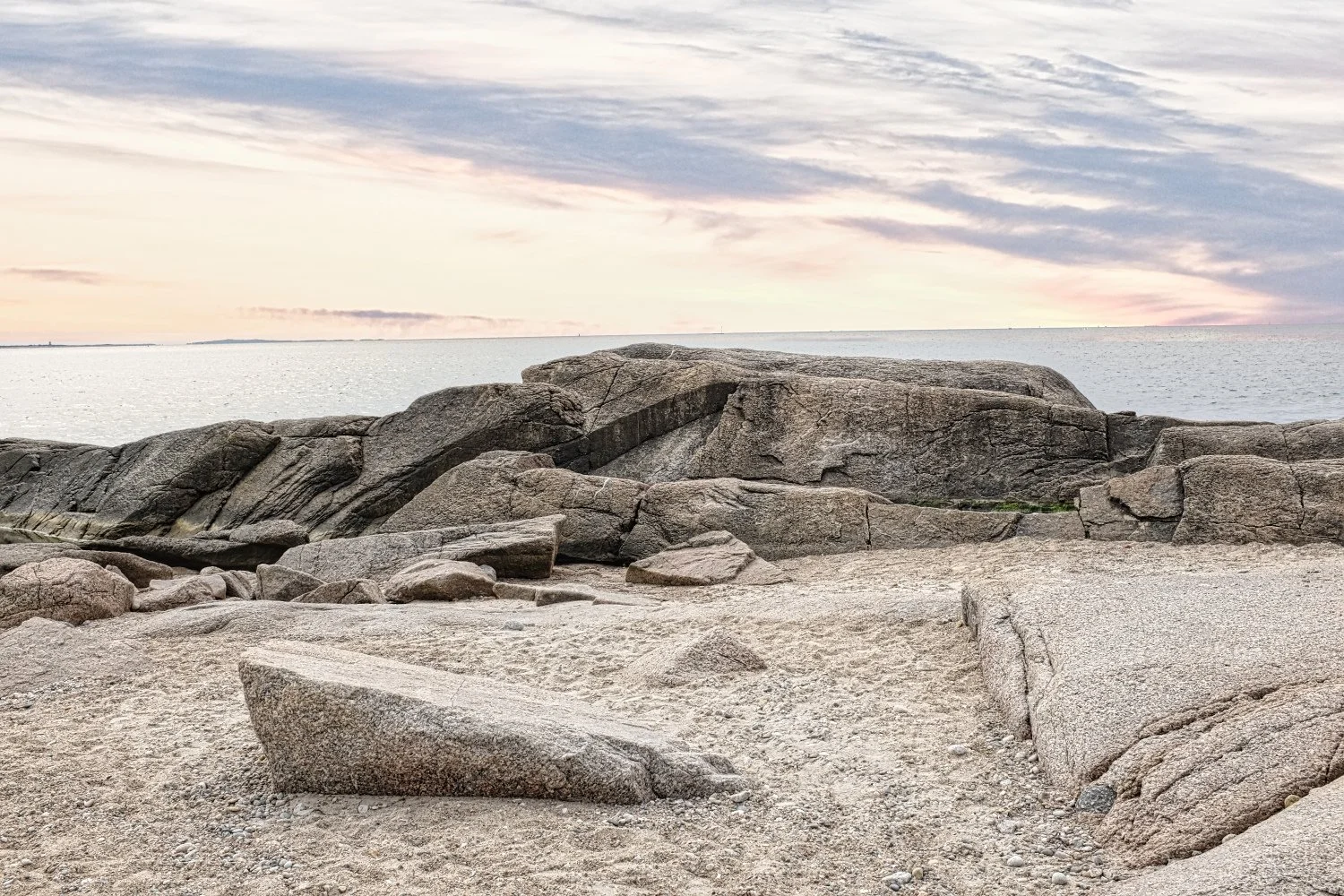 A rocky beach scene at sunset with large granite boulders and a calm ocean in the background. The sky has pastel-colored clouds.