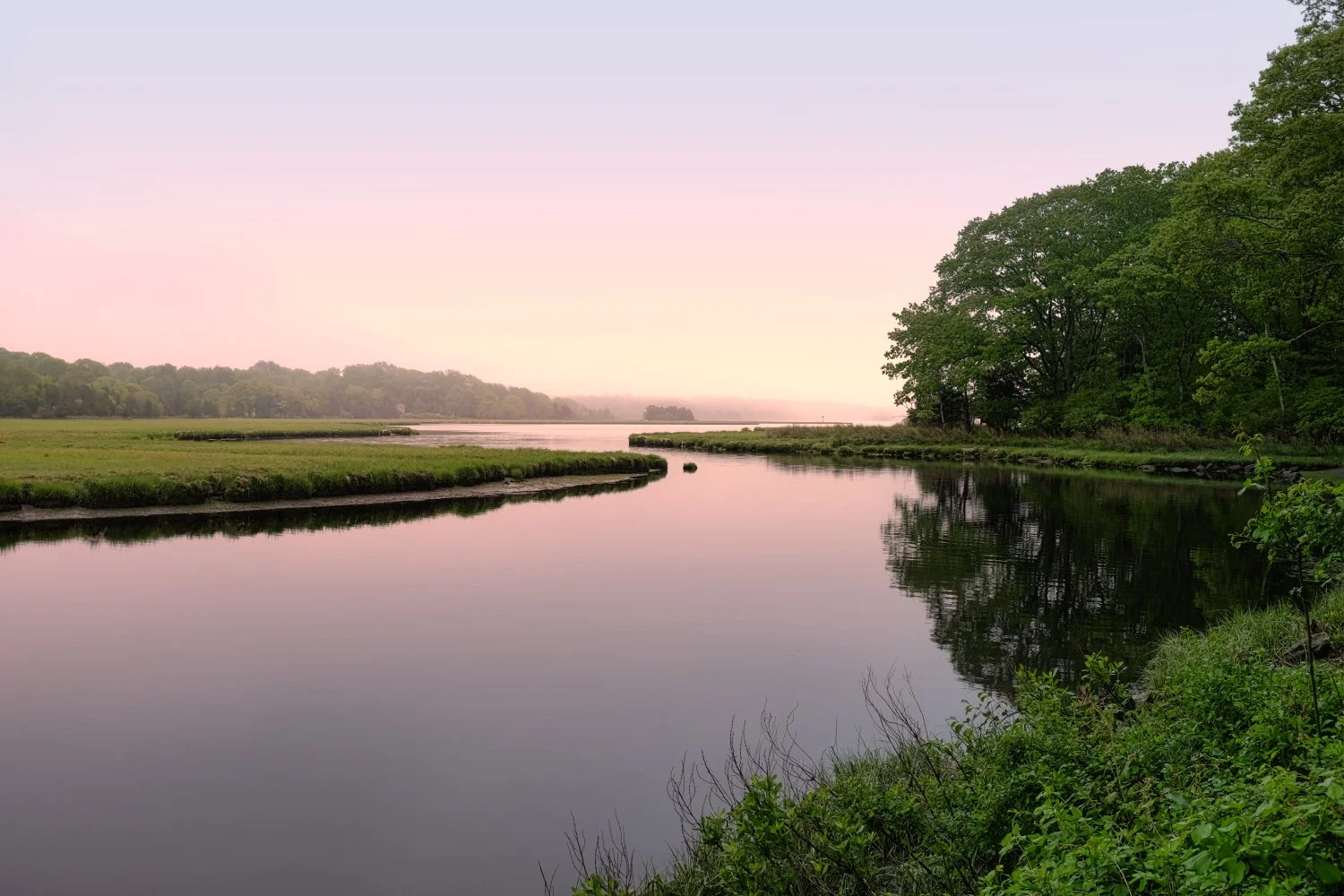 A calm river reflecting the green trees and pink sky during sunrise or sunset, with grassy banks and distant hills.