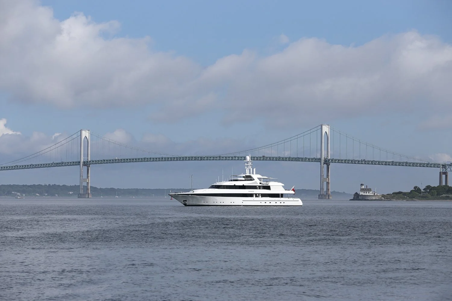 A large white yacht sailing on calm water with a bridge in the background and partly cloudy sky.