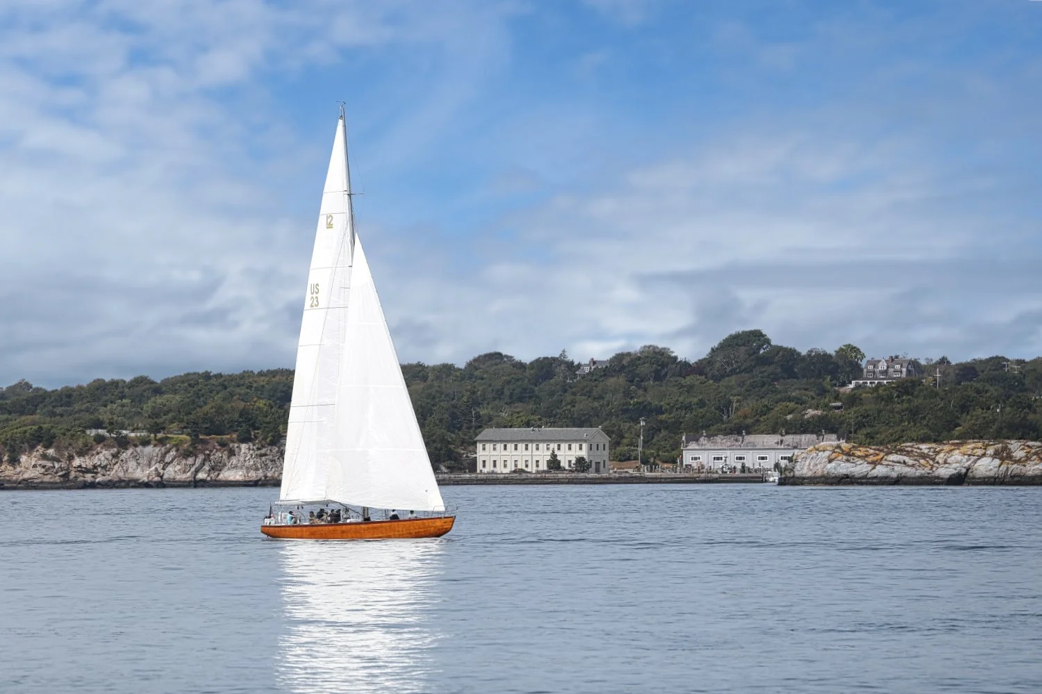 A sailboat with a white sail and wooden hull sailing on calm water near a rocky shoreline with houses and green trees in the background under a partly cloudy sky.