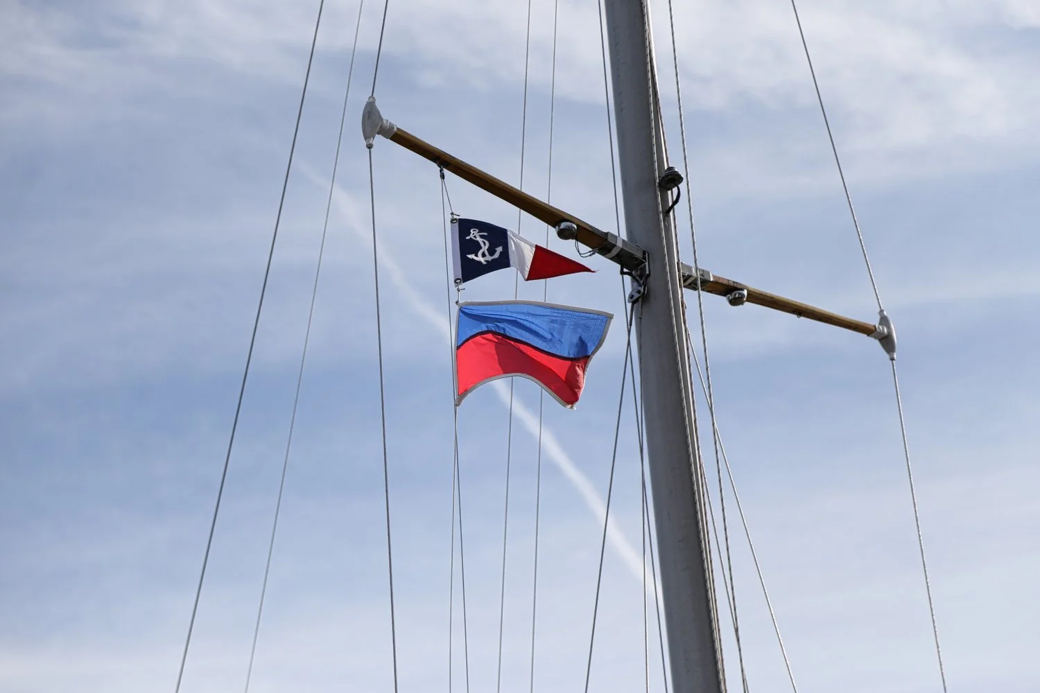 Three flags on a sailboat mast against a blue sky with wispy clouds, including a Nautical flag, a French flag, and a red and blue flag.