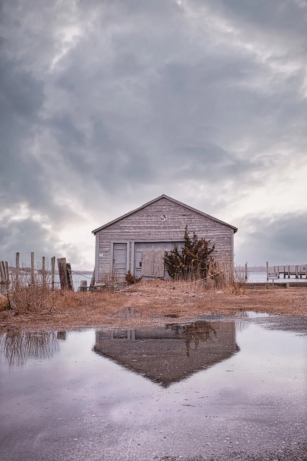 A weathered gray wooden barn with boarded-up doors and windows, surrounded by dry grass and bushes, under a cloudy sky, with its reflection visible in a large puddle in the foreground.