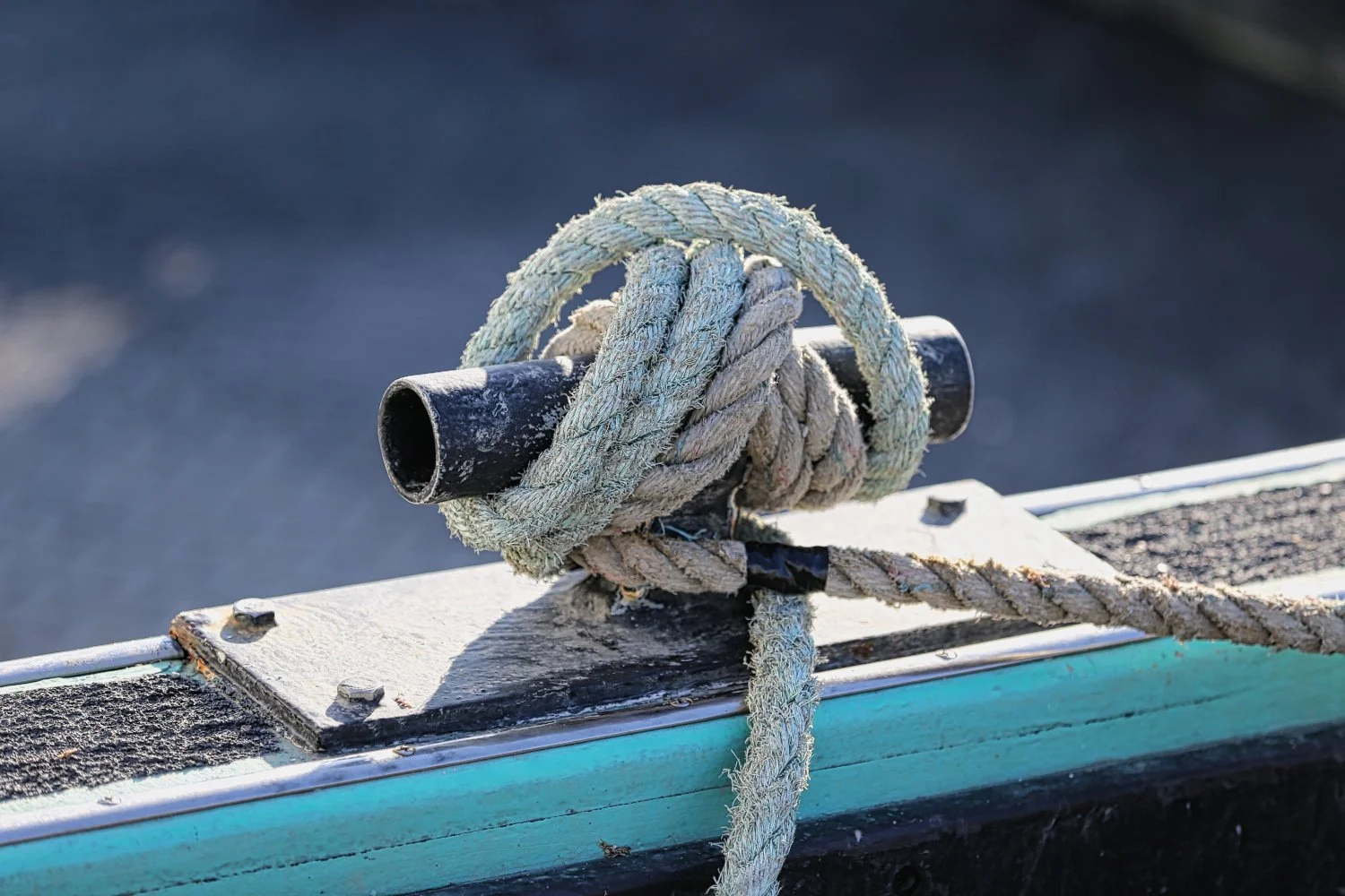Close-up of a boat's metal cleat with a thick, weathered rope tied around it, on a dock or boat deck.