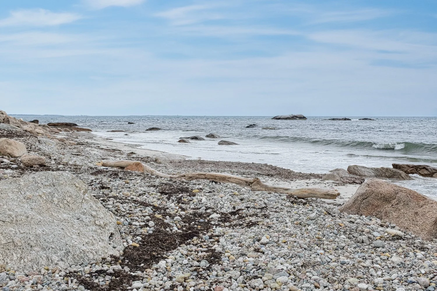A rocky beach with pebbles and driftwood, ocean waves, and a partly cloudy sky.