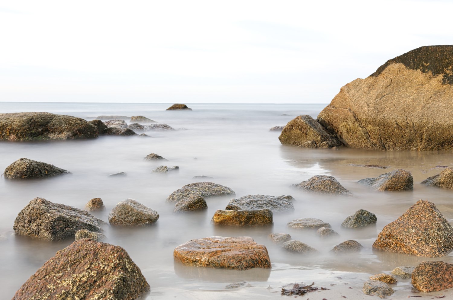 Calm ocean water with large rocks and smaller stones on the sandy shore, some partially submerged, under a light sky.
