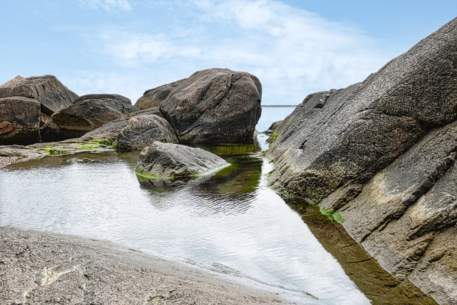 Rocks and boulders along the shoreline with shallow water reflecting the sky, some green moss on the rocks, and a distant view of the water and sky at the horizon.