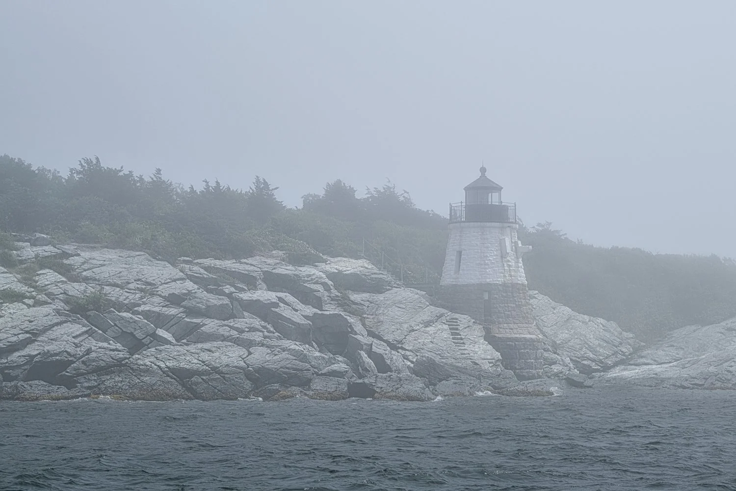 A foggy scene of a lighthouse on a rocky shoreline, with dense vegetation and trees in the background, water in the foreground, and a misty atmosphere.