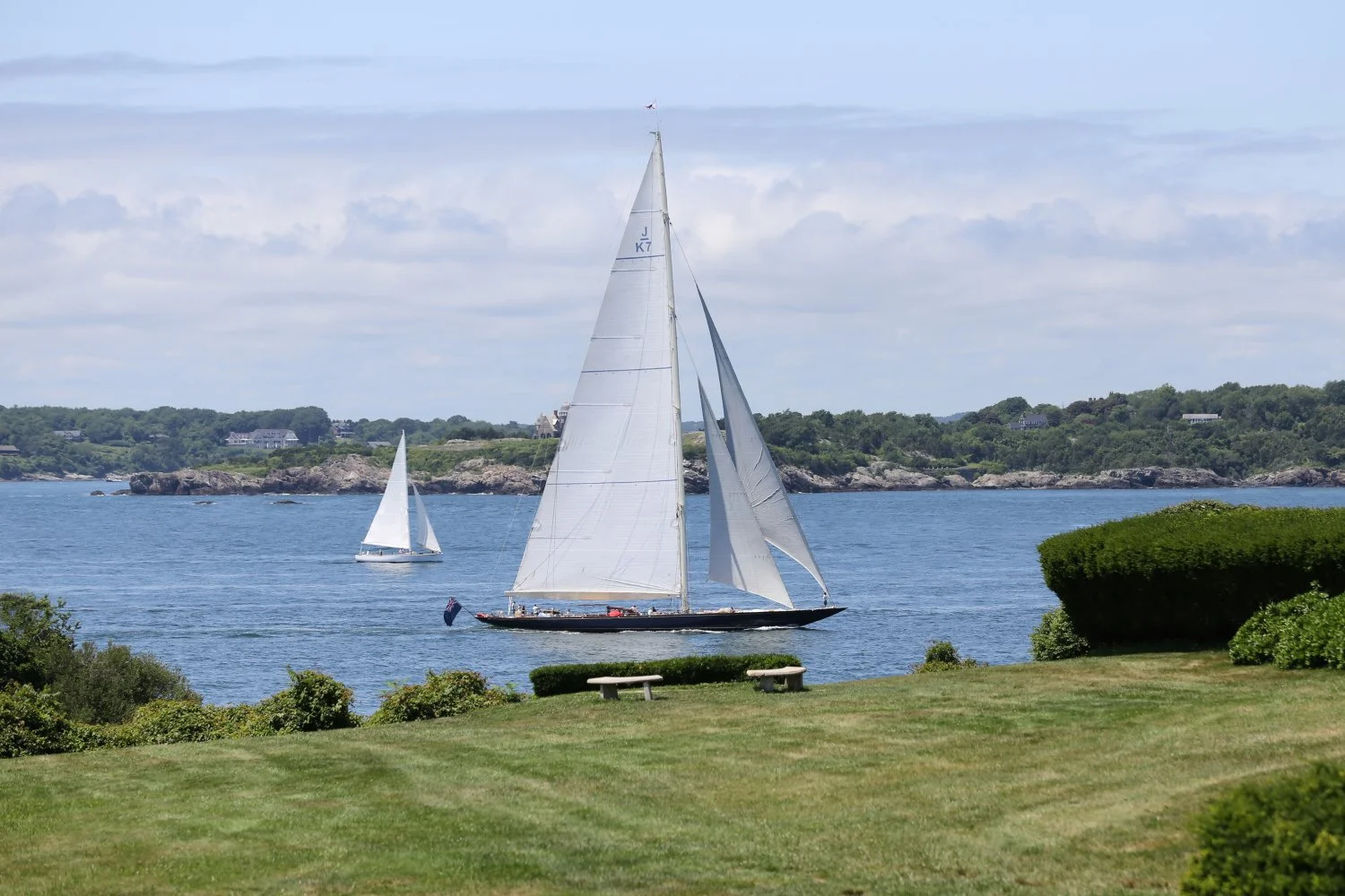 A sailboat on the water with its sails up, with another smaller sailboat in the background, near a grassy shoreline with benches and bushes, and a distant tree-lined coast under a partly cloudy sky.