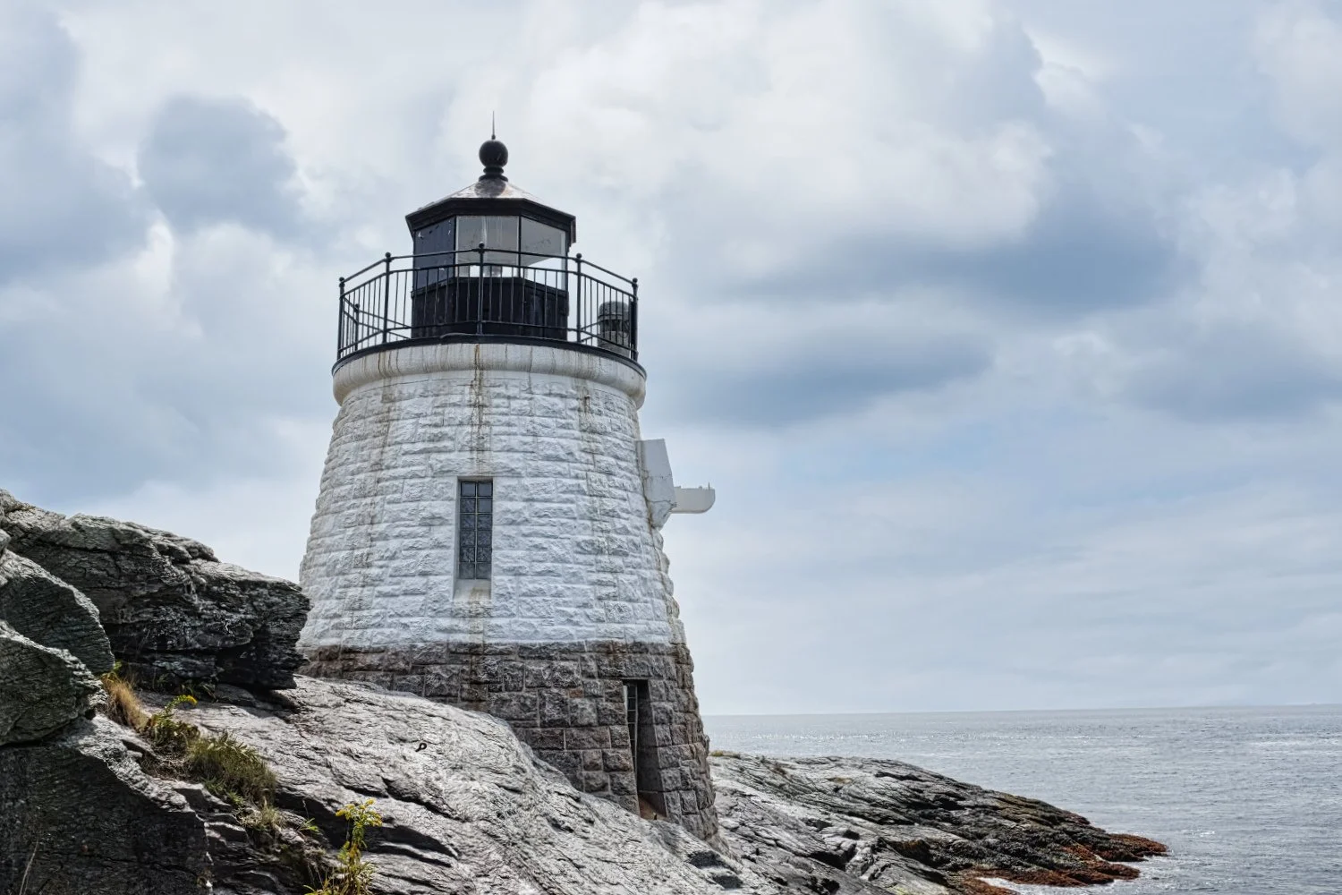 White lighthouse on rocky coast under cloudy sky with ocean in the background.