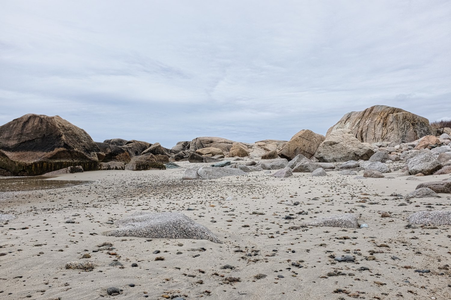 A sandy beach with large rocks and boulders under an overcast sky.