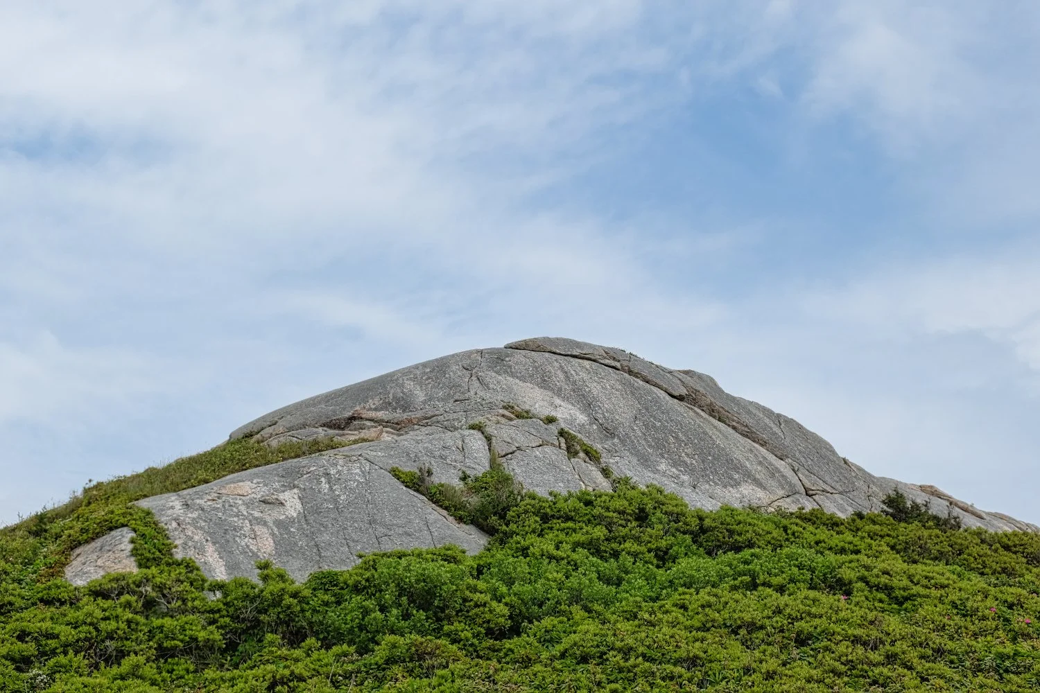 Large gray granite hill with green shrubs at the base and a partly cloudy blue sky