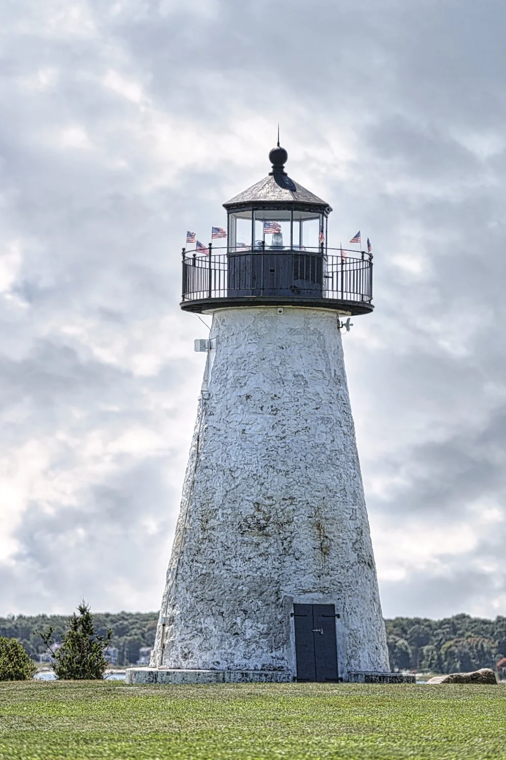 A white lighthouse with a black lantern room and a railing on top, decorated with several small American flags, standing on a grassy area under a cloudy sky.