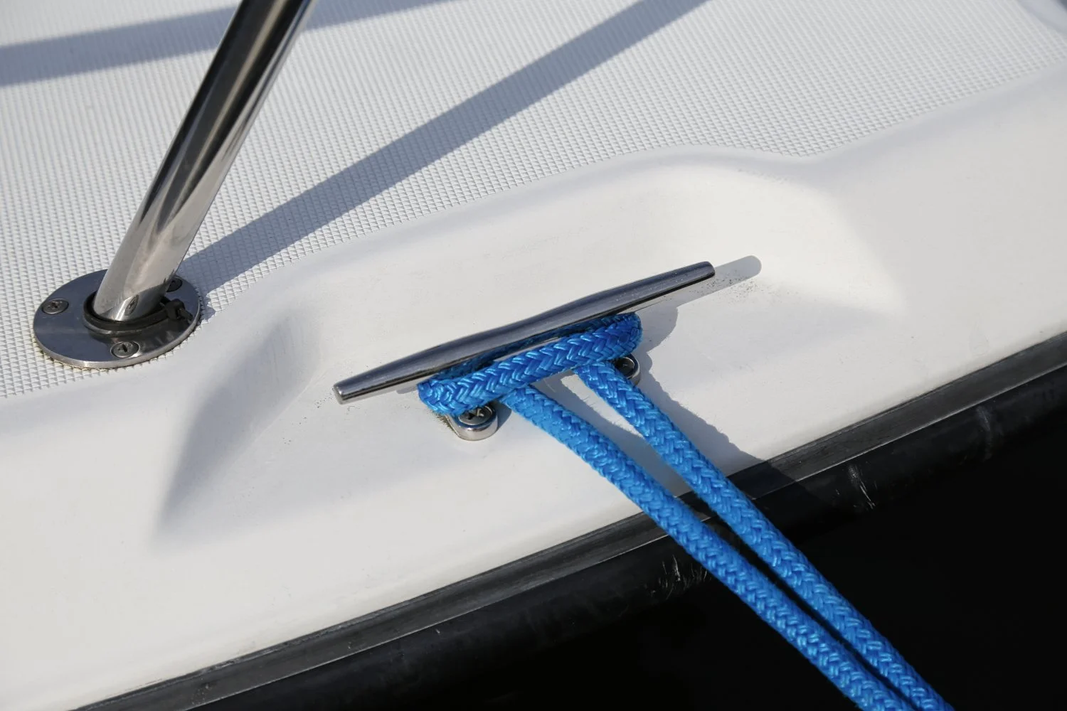 Close-up of a boat's cleat with a blue rope tied around it, mounted on a white deck with metal hardware and a stainless-steel support structure.