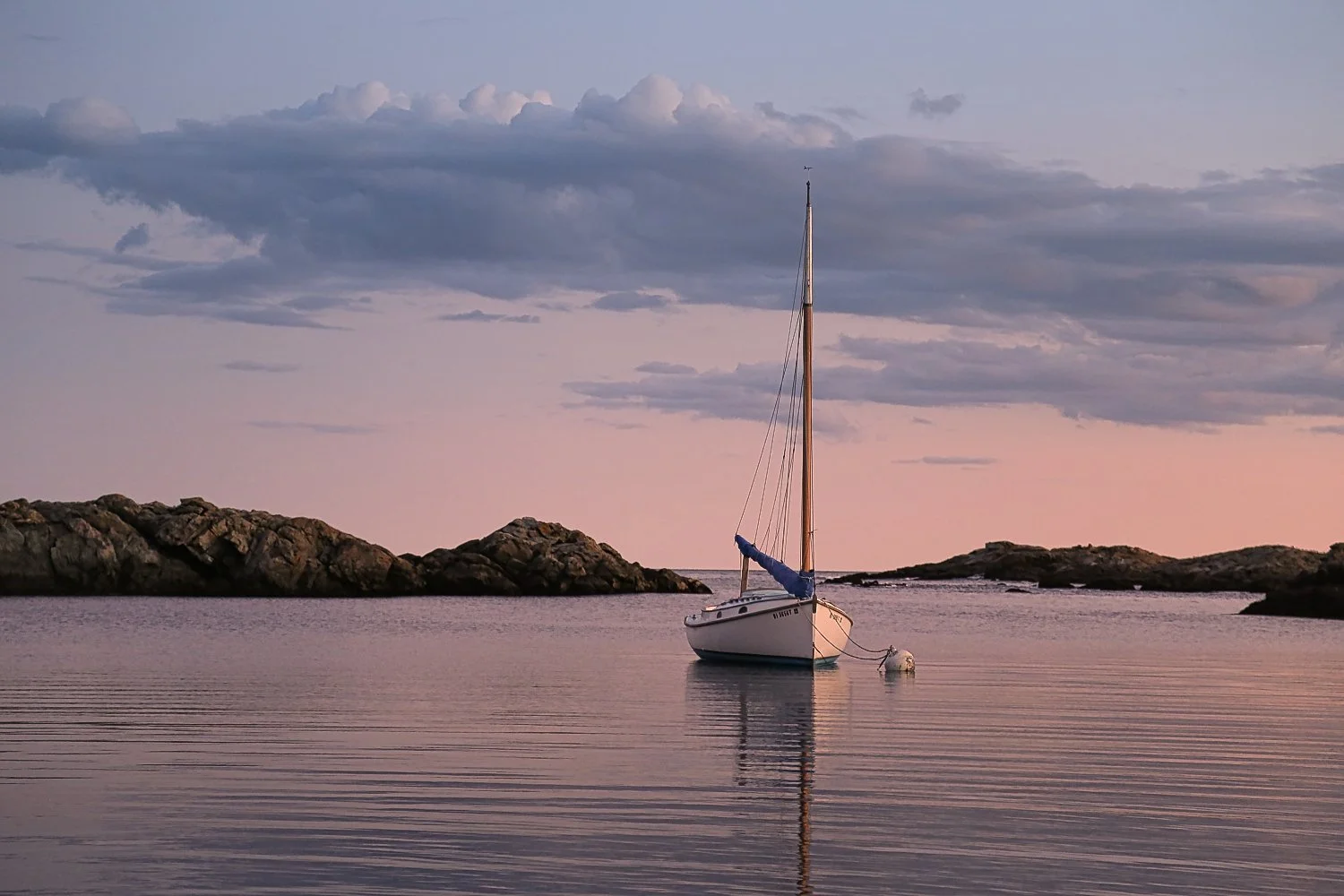 A sailboat anchored in calm water near rocky islands during sunset or sunrise, with pastel-colored sky and clouds.
