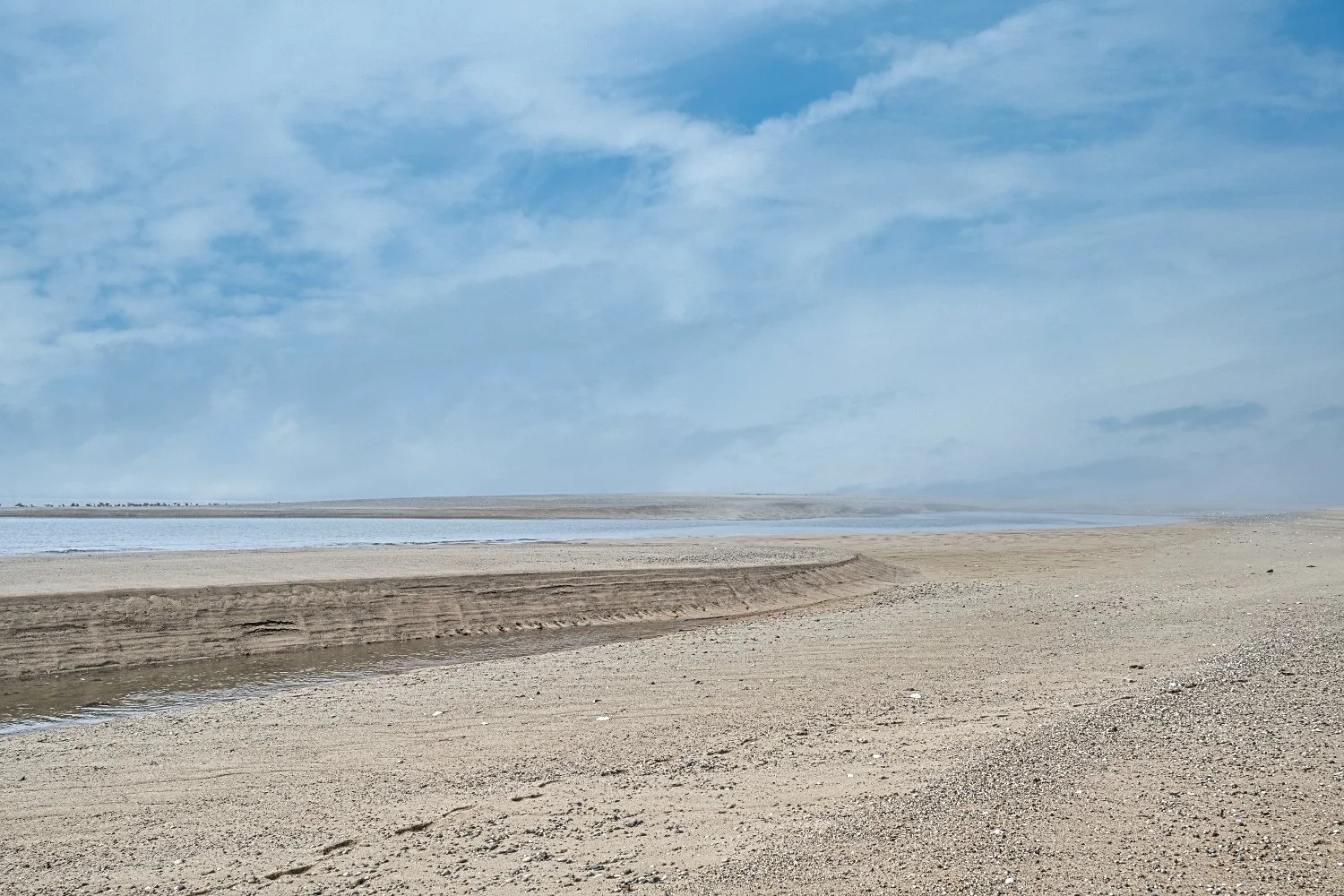 Expansive sandy beach with a waterway or lagoon, under a partly cloudy sky