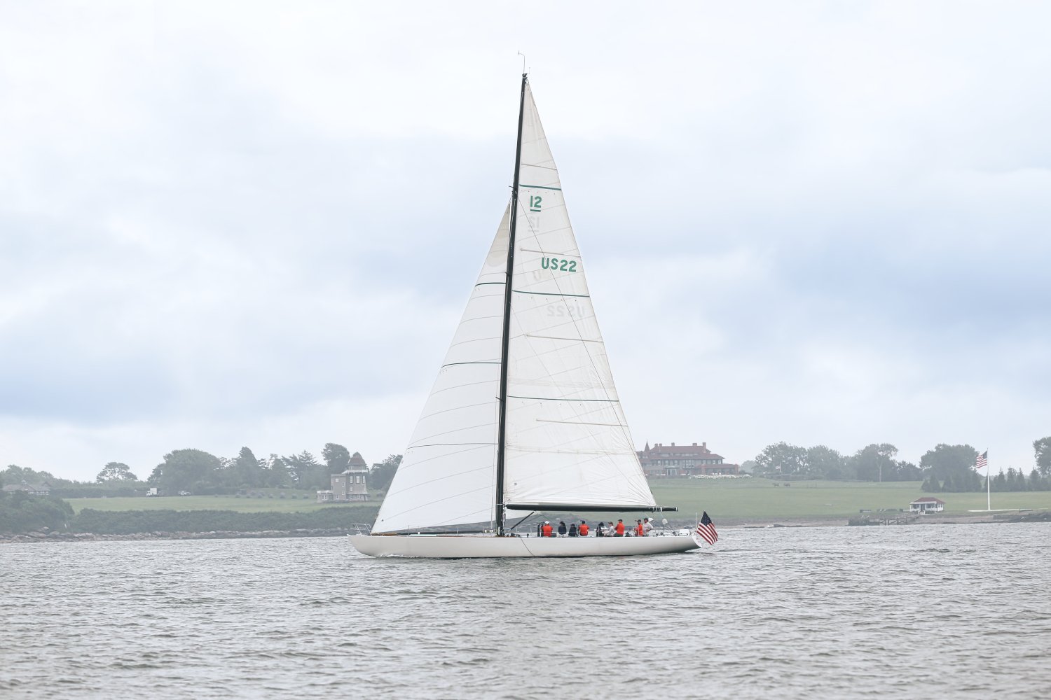A sailboat with white sails on the water, with a group of people on board, and an American flag at the stern. In the background, there are houses and trees along the shoreline, under a cloudy sky.