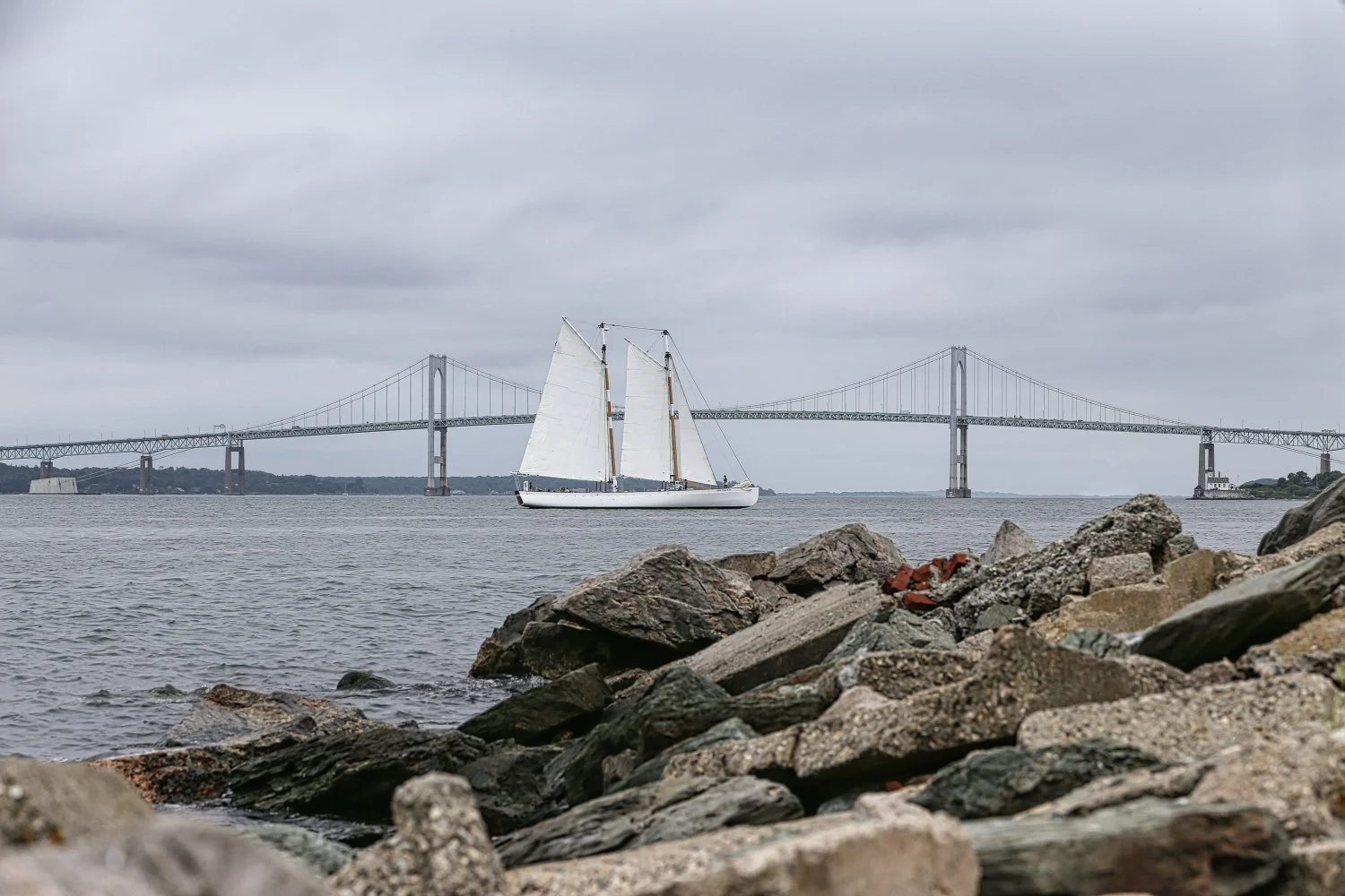 A sailboat with white sails on a body of water near a rocky shoreline, with a large suspension bridge and cloudy sky in the background.