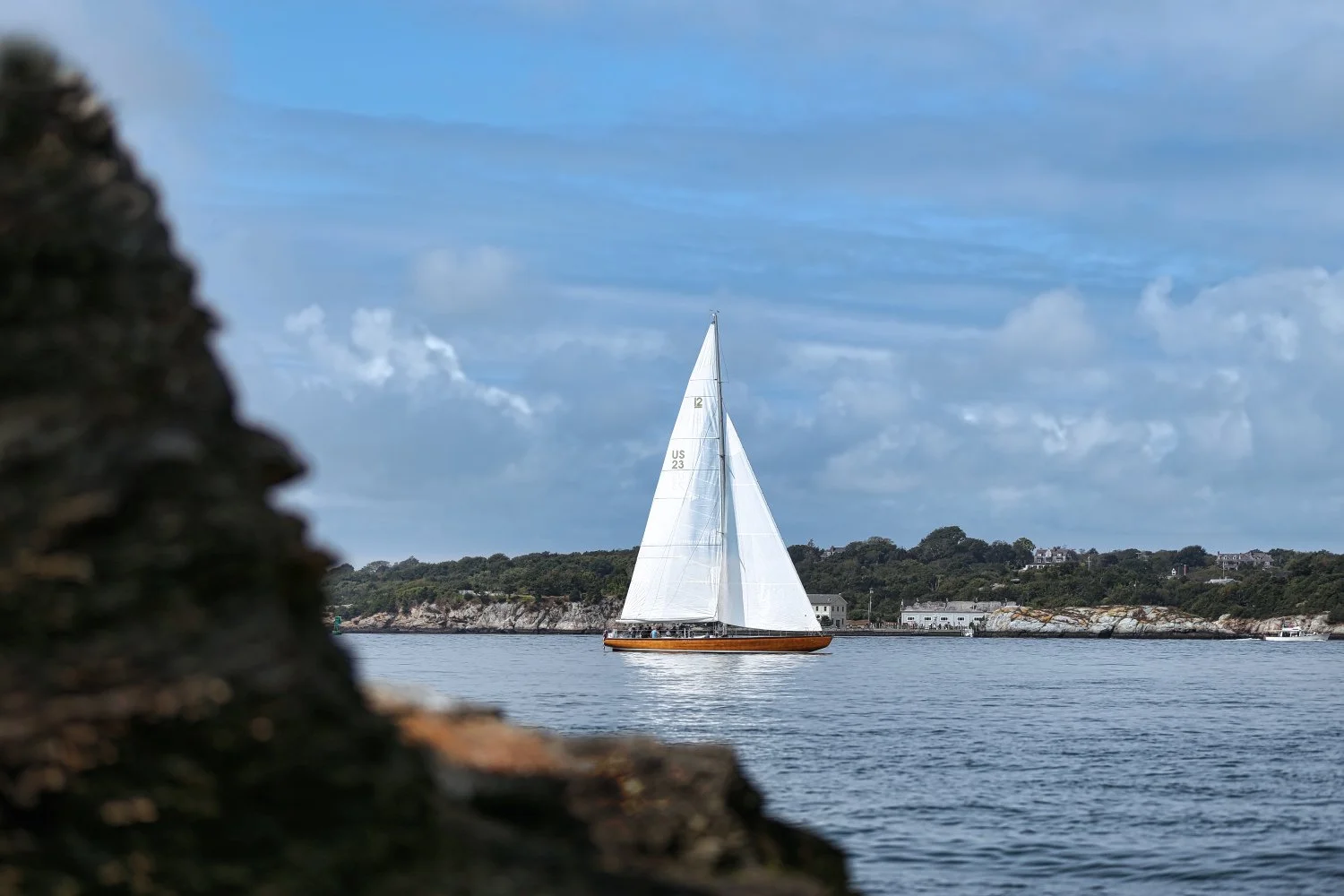 A sailboat with white sails on calm water, with a rocky shoreline and trees in the background under a partly cloudy sky.