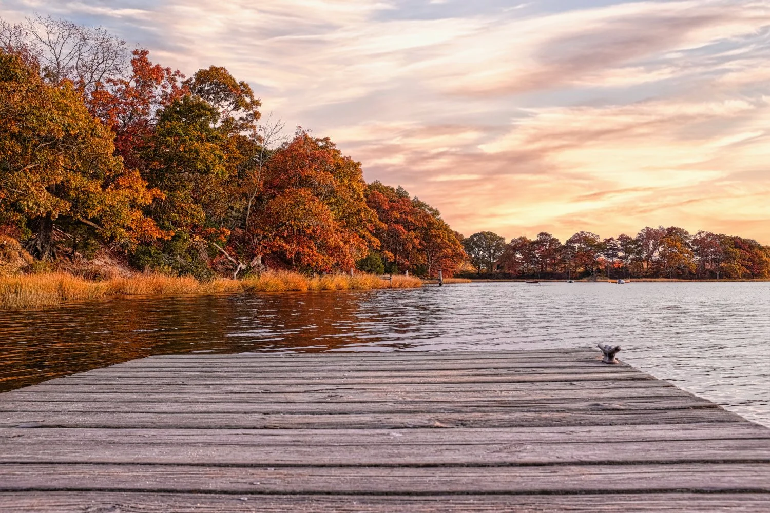 A wooden dock extends into a calm lake during sunset, with trees in fall colors along the shoreline and a pink and orange sky.