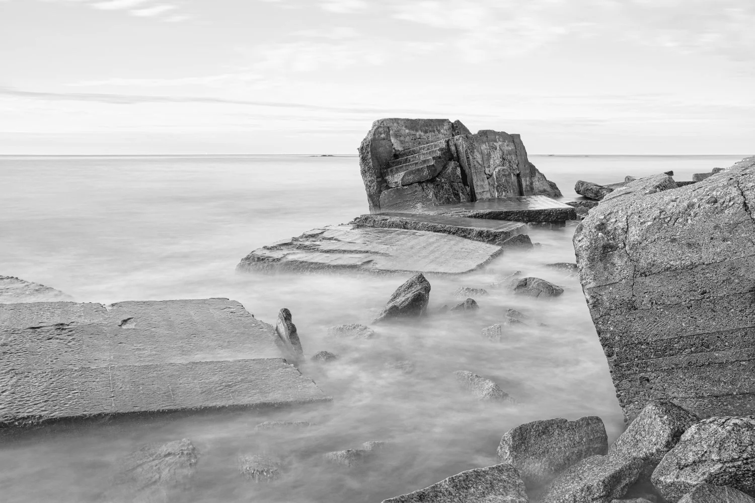 Black-and-white photo of large rocks and concrete blocks near the shoreline with water gently flowing around them, and a cloudy sky in the background.