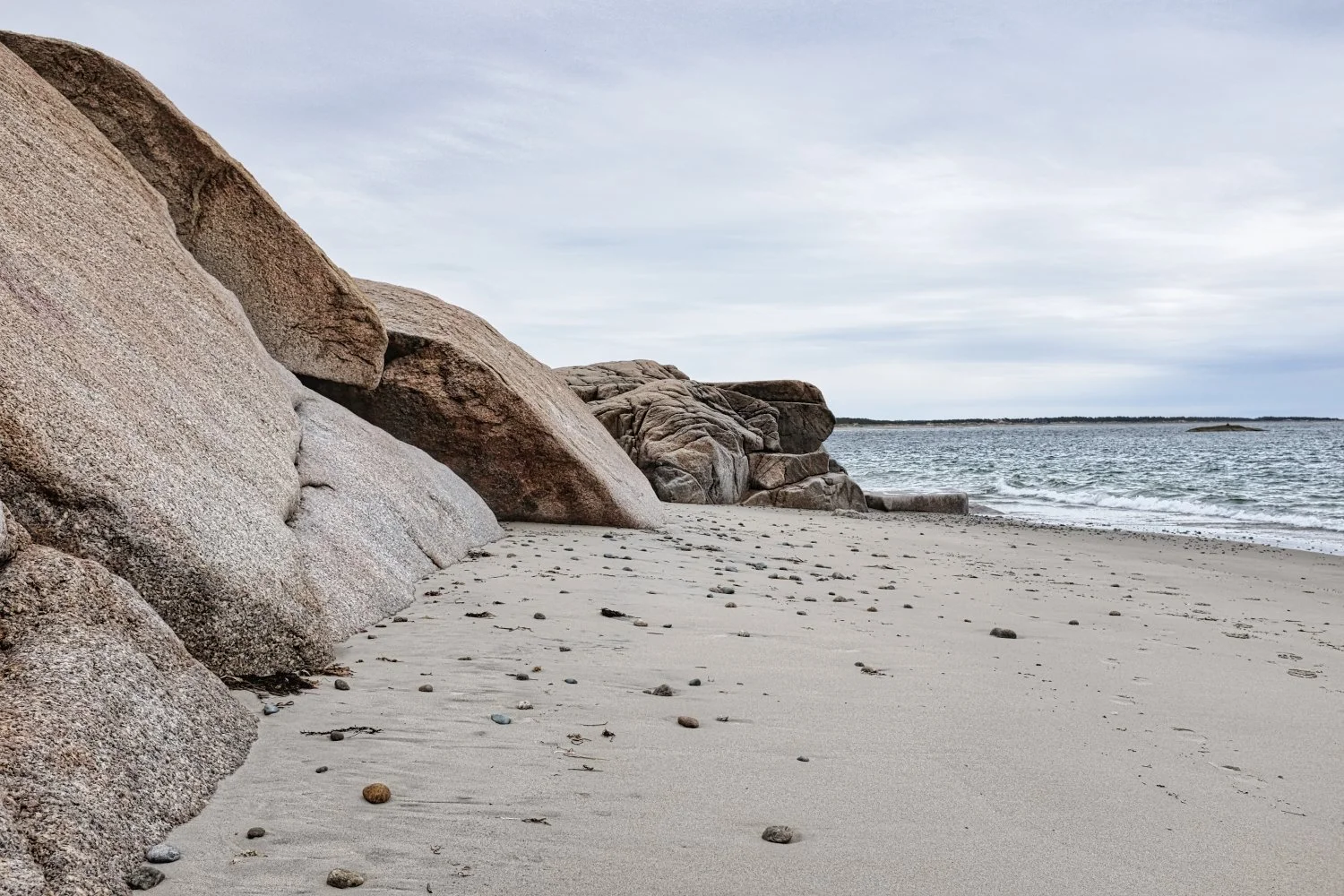 A sandy beach with large granite rocks on the left, scattered pebbles on the sand, and the ocean with small waves to the right with an overcast sky in the background.