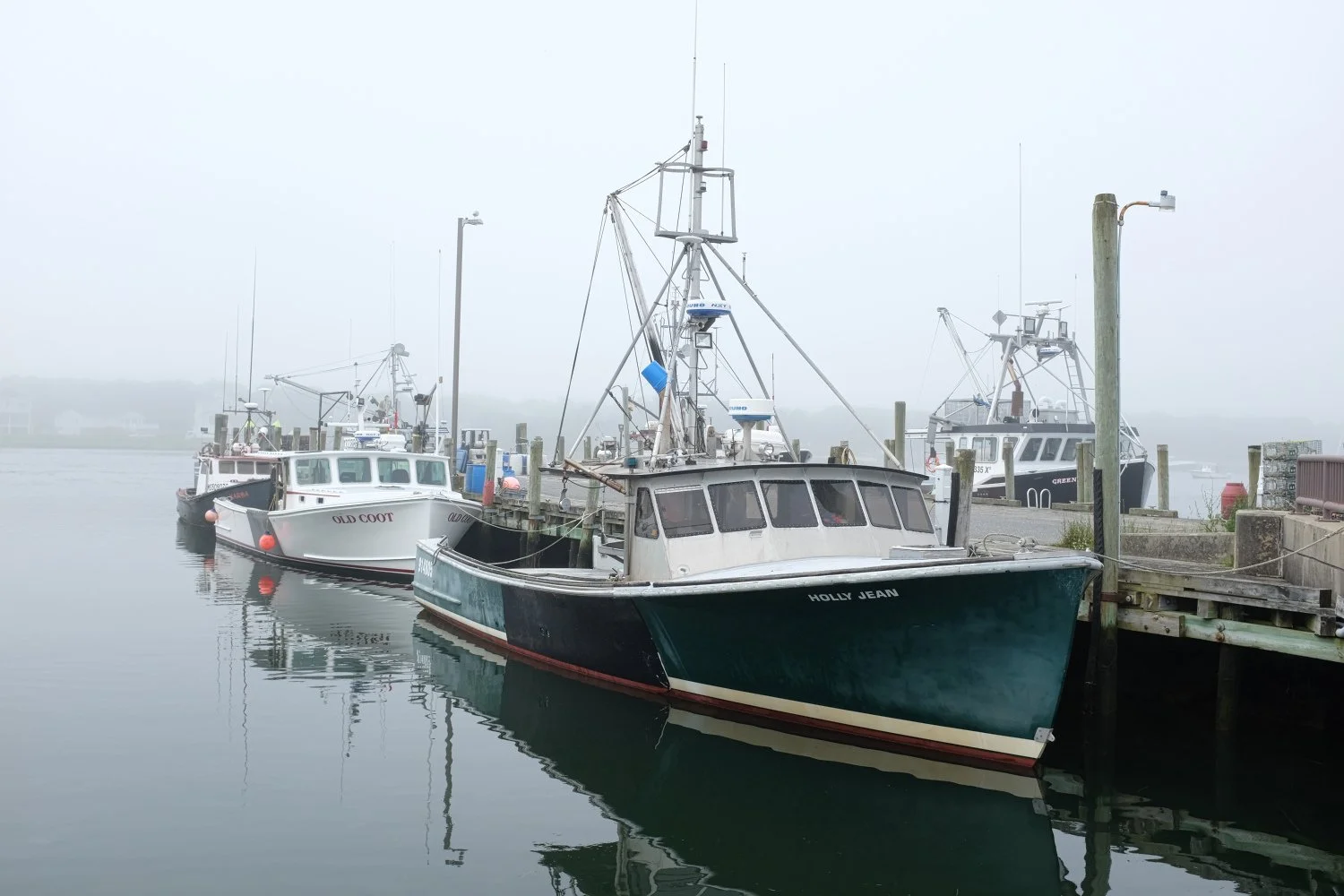 Several boats docked at a foggy marina, including one named 'Holly Jean' in the foreground and others like 'Old Coot' beside it.