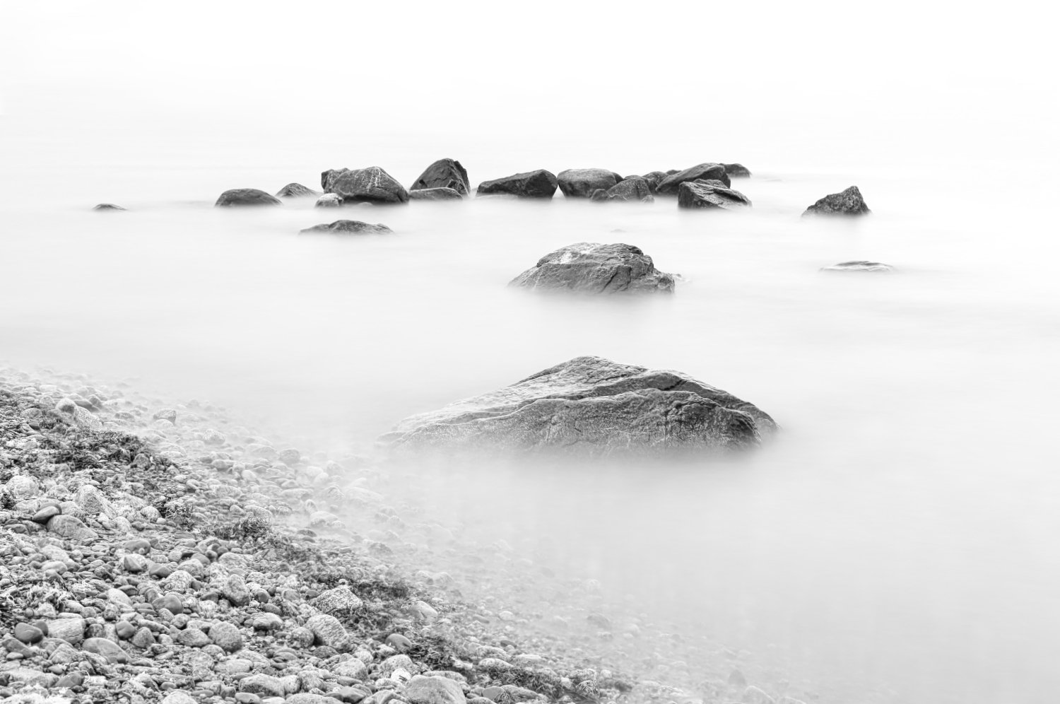 Black and white photo of rocks in calm water near a pebbled shoreline.