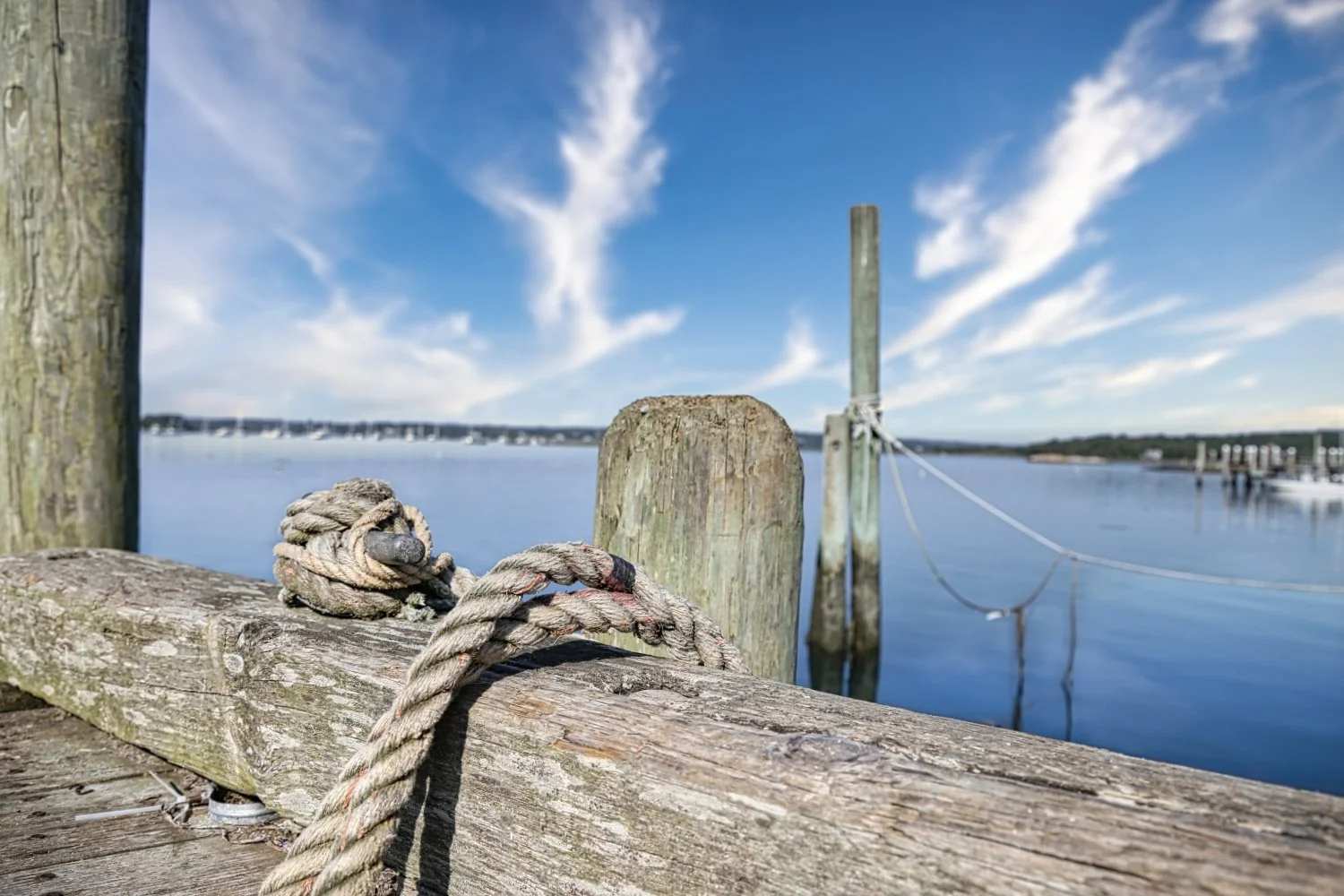 Close-up of a weathered nautical rope tied around a wooden dock post, with a calm blue water body and boats in the distance under a partly cloudy sky.
