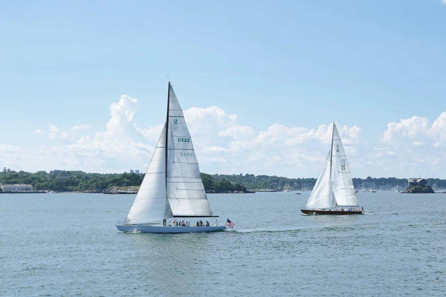 Two sailboats on a calm body of water with a distant shoreline and partly cloudy sky
