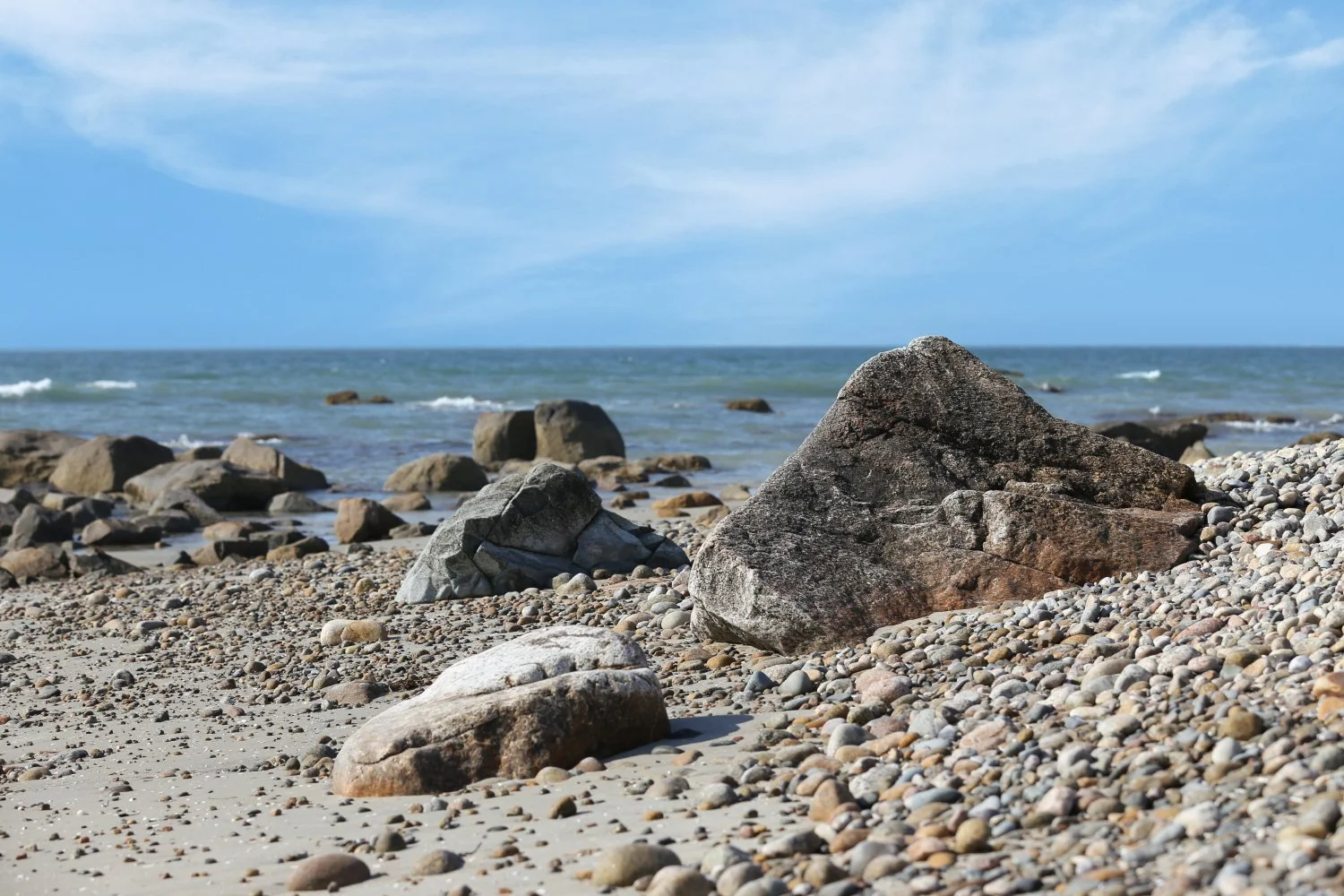 Rocky beach with large and small stones, ocean waves, and blue sky with some clouds.