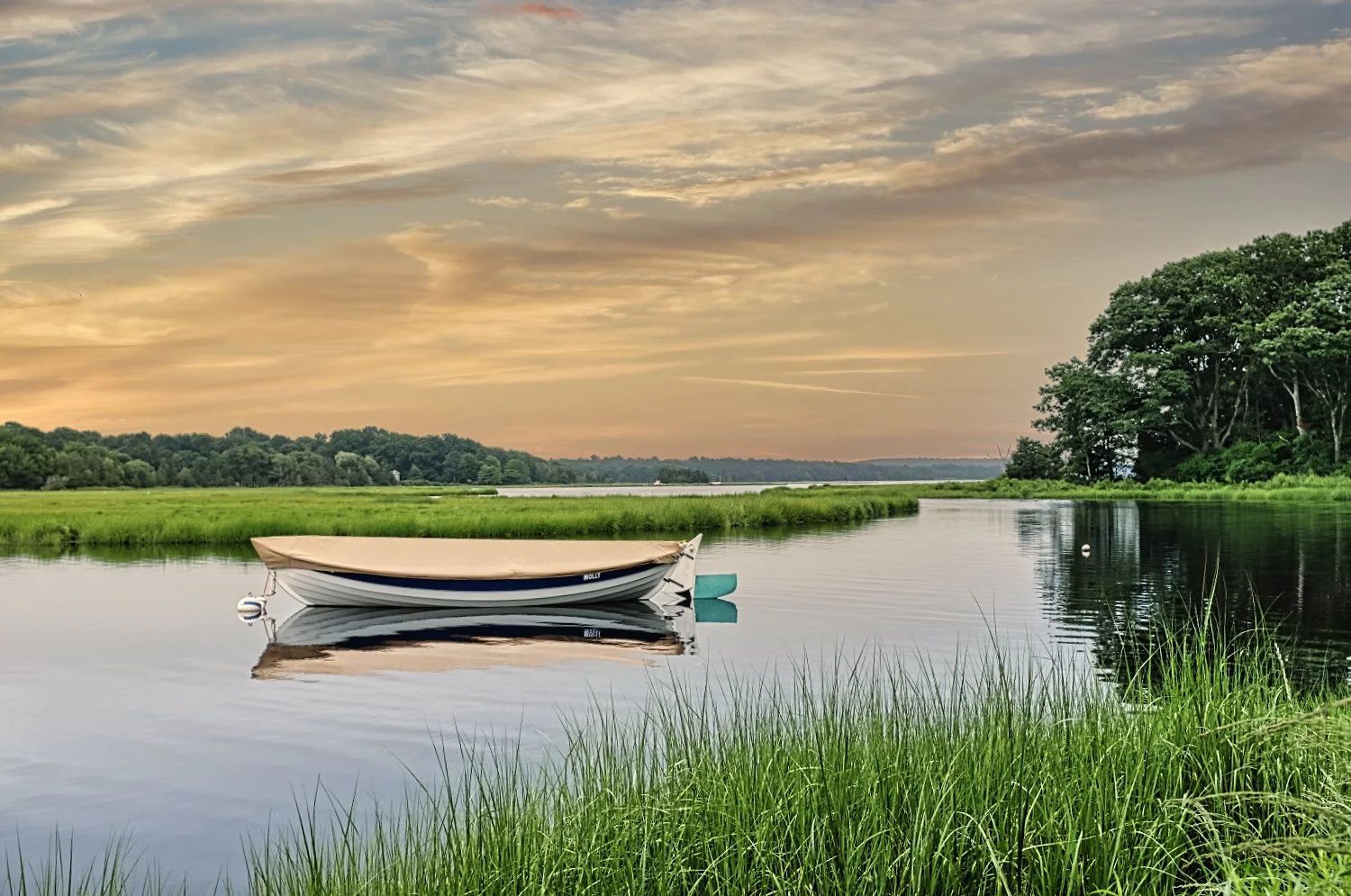 A small boat floating on calm river water with green grass in the foreground and a tree-lined bank in the background during sunset.