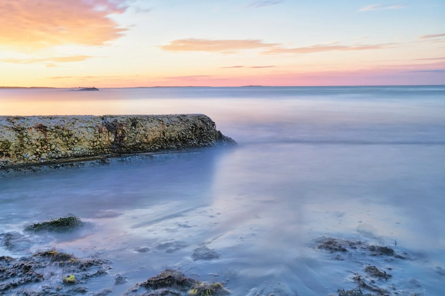A seascape at sunset with a rocky shoreline, a weathered concrete pier, calm water, and a colorful sky with pink, orange, and purple hues.