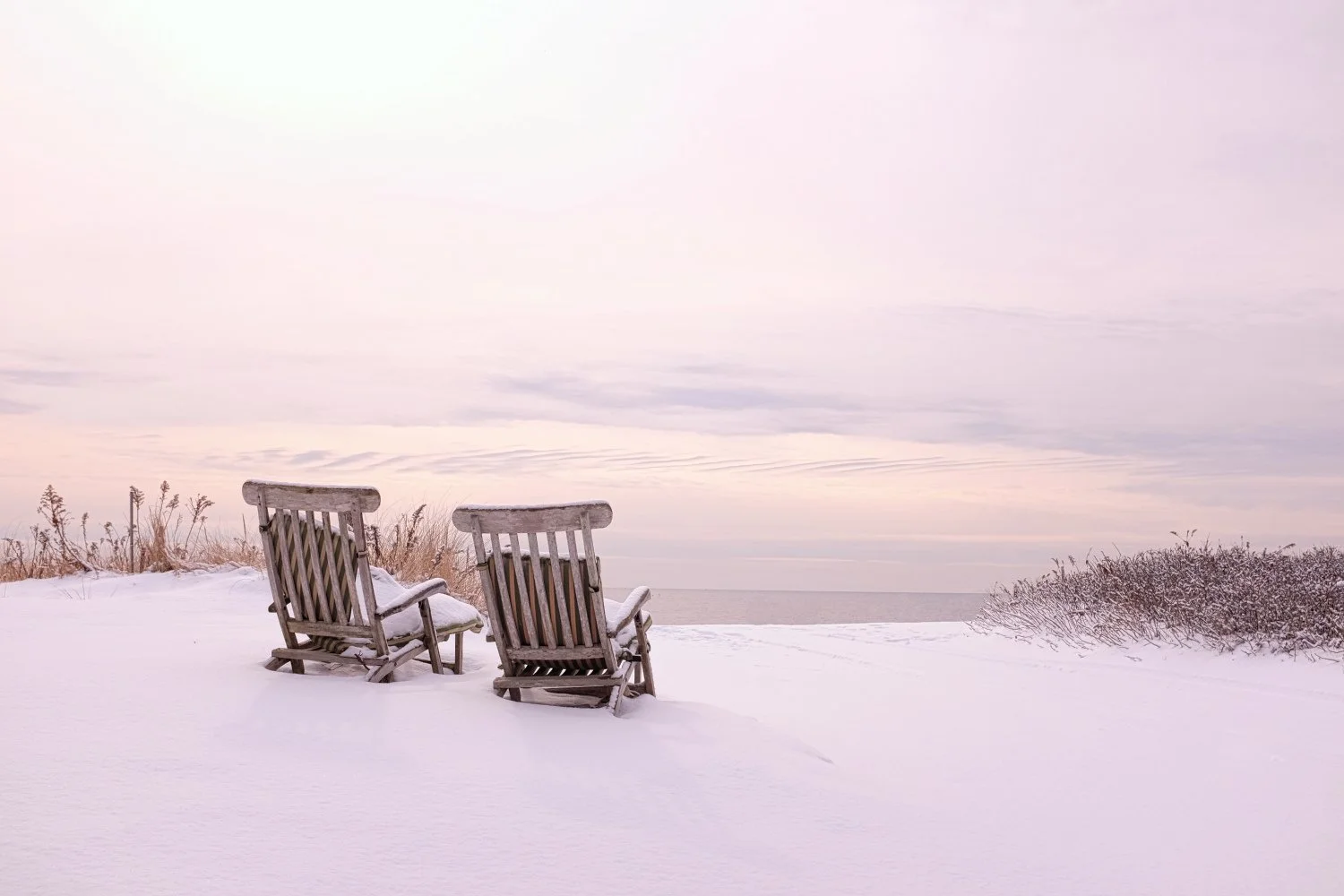 Two weathered wooden chairs covered in snow facing a snow-covered beach and ocean under a cloudy sky with soft pastel hues.