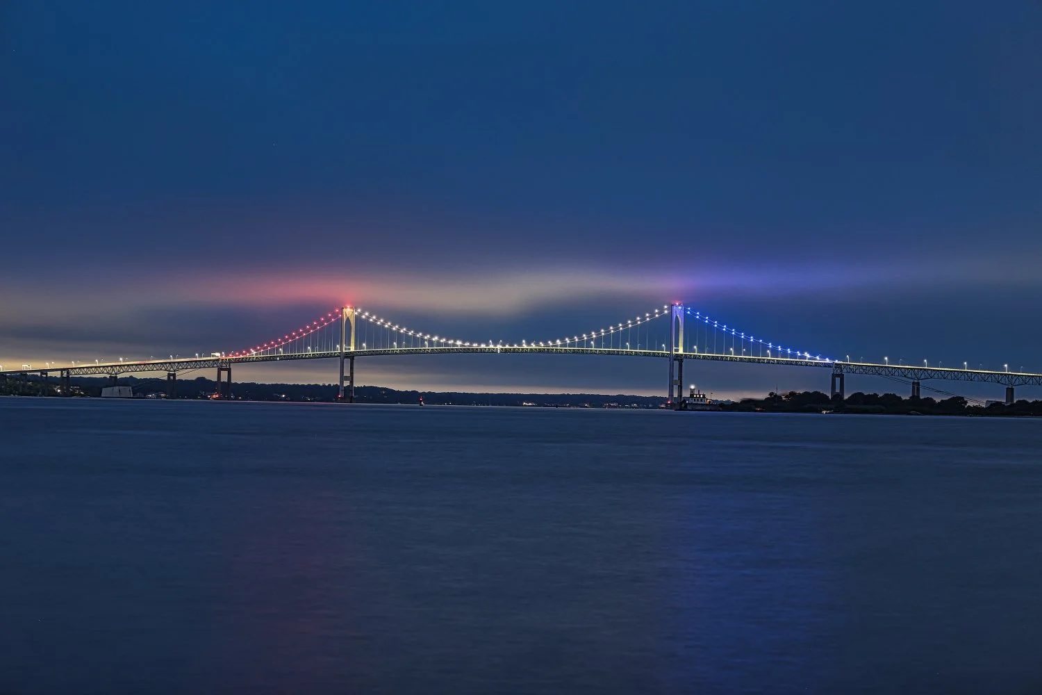 Nighttime view of a large suspension bridge illuminated with red, white, and blue lights, spanning across a body of water under a cloudy sky.