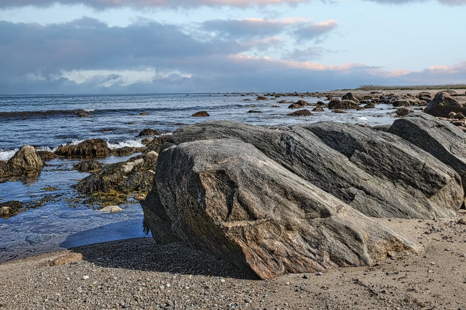 Beach scene with large rocks on sandy shore, calm ocean water, and a cloudy sky.
