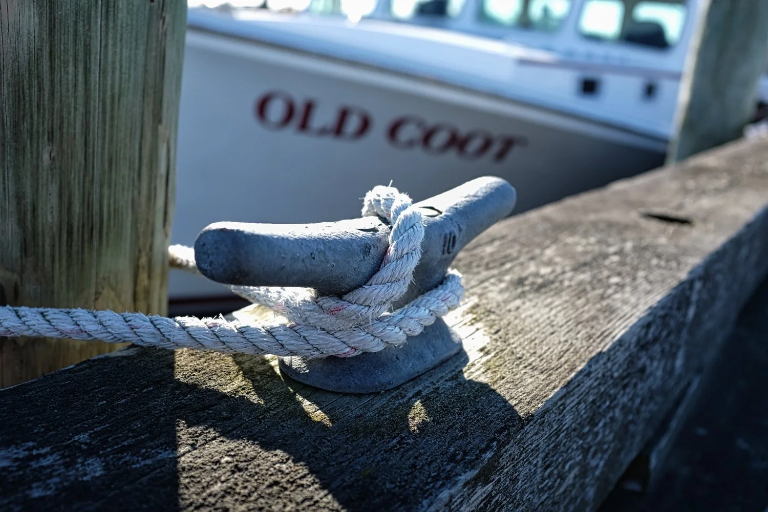 Close-up of a boat mooring cleat with a white rope wrapped around it, attached to a weathered wooden post, with a boat labeled 'OLD COOT' in the background.