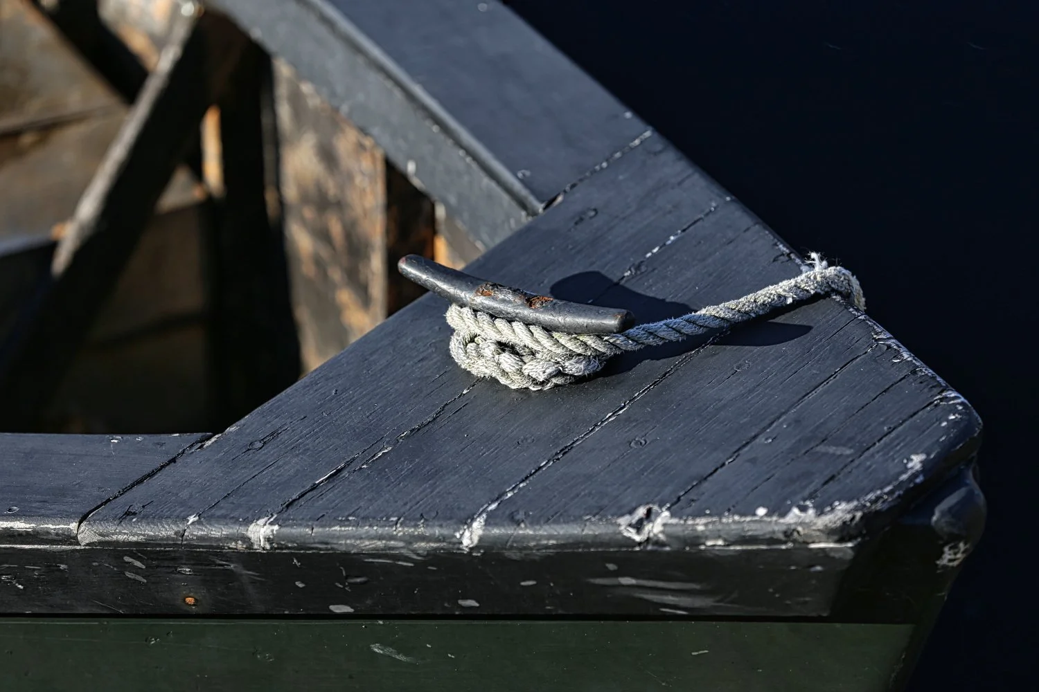 Close-up of a metal boat cleat with a looped rope tied around it on a black painted boat. The boat's wooden surface shows signs of wear and chipping.