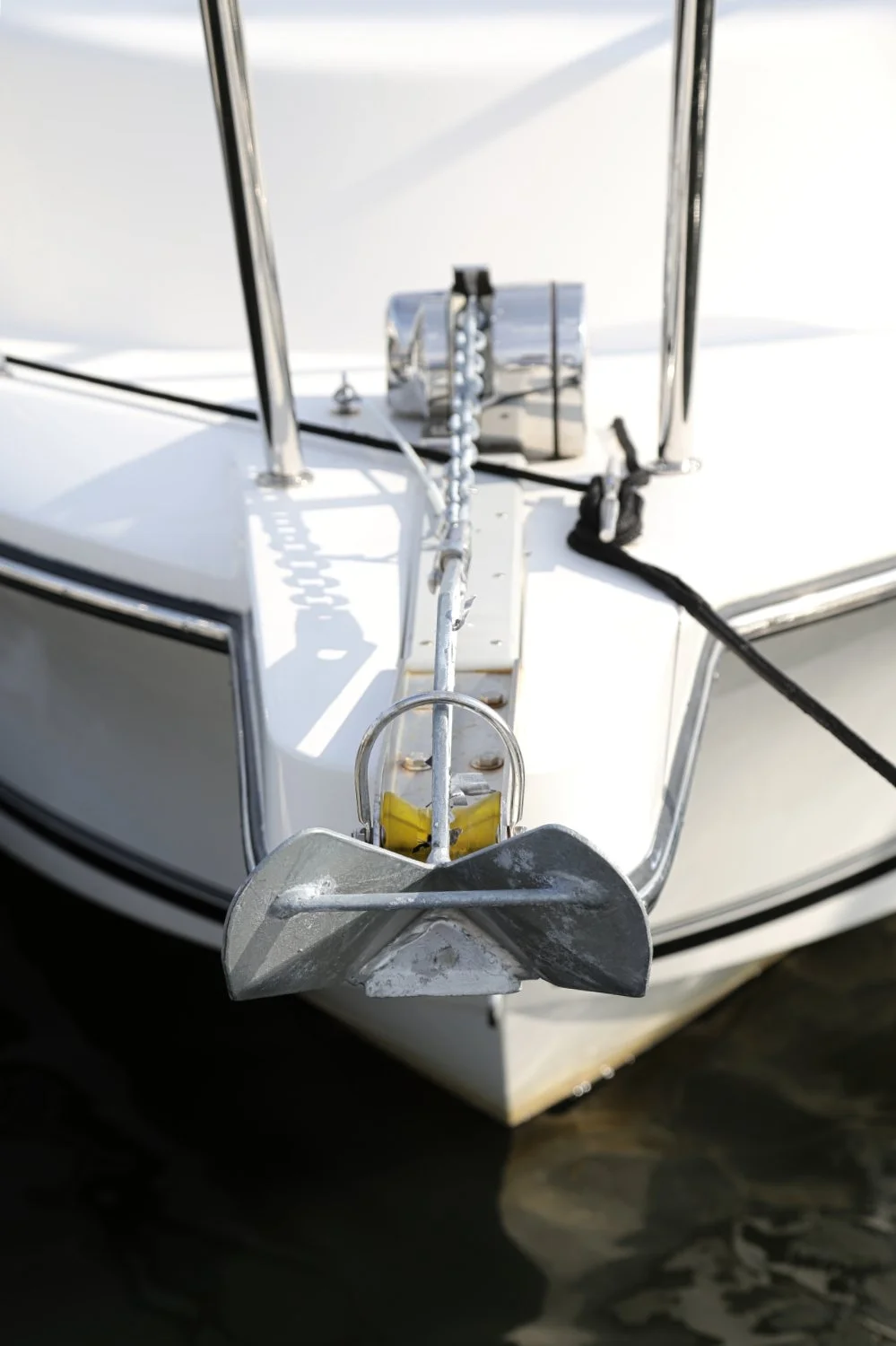 Close-up of a boat's bow with an anchor and chain secured to the deck, with a chrome fitting and black rope, and the boat floating on water.