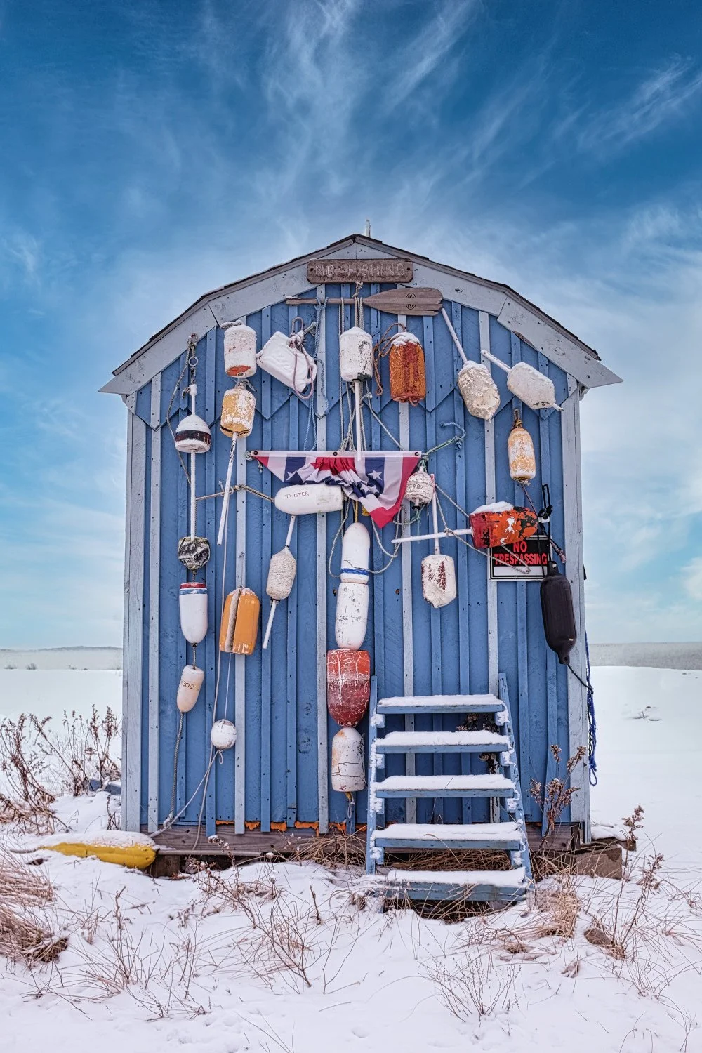 A blue beach hut with a collection of old buoys and floats hanging on the front, a small set of snow-covered stairs, and a partially visible 