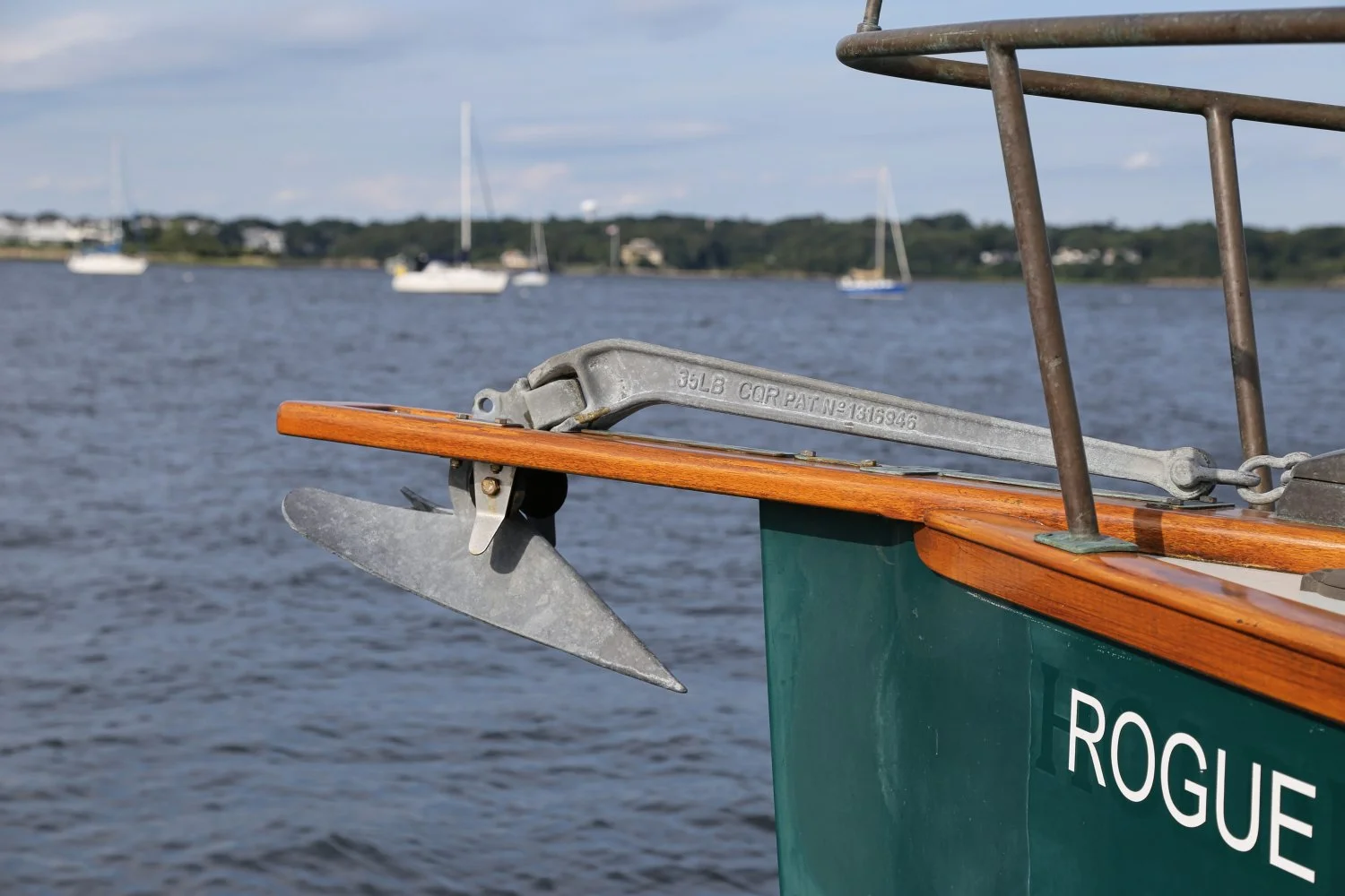 Close-up of a boat's bow with an anchor secured to the front, displaying the boat name 'Rogue' on the side. The background shows a body of water with several sailboats and a distant shoreline under a partly cloudy sky.