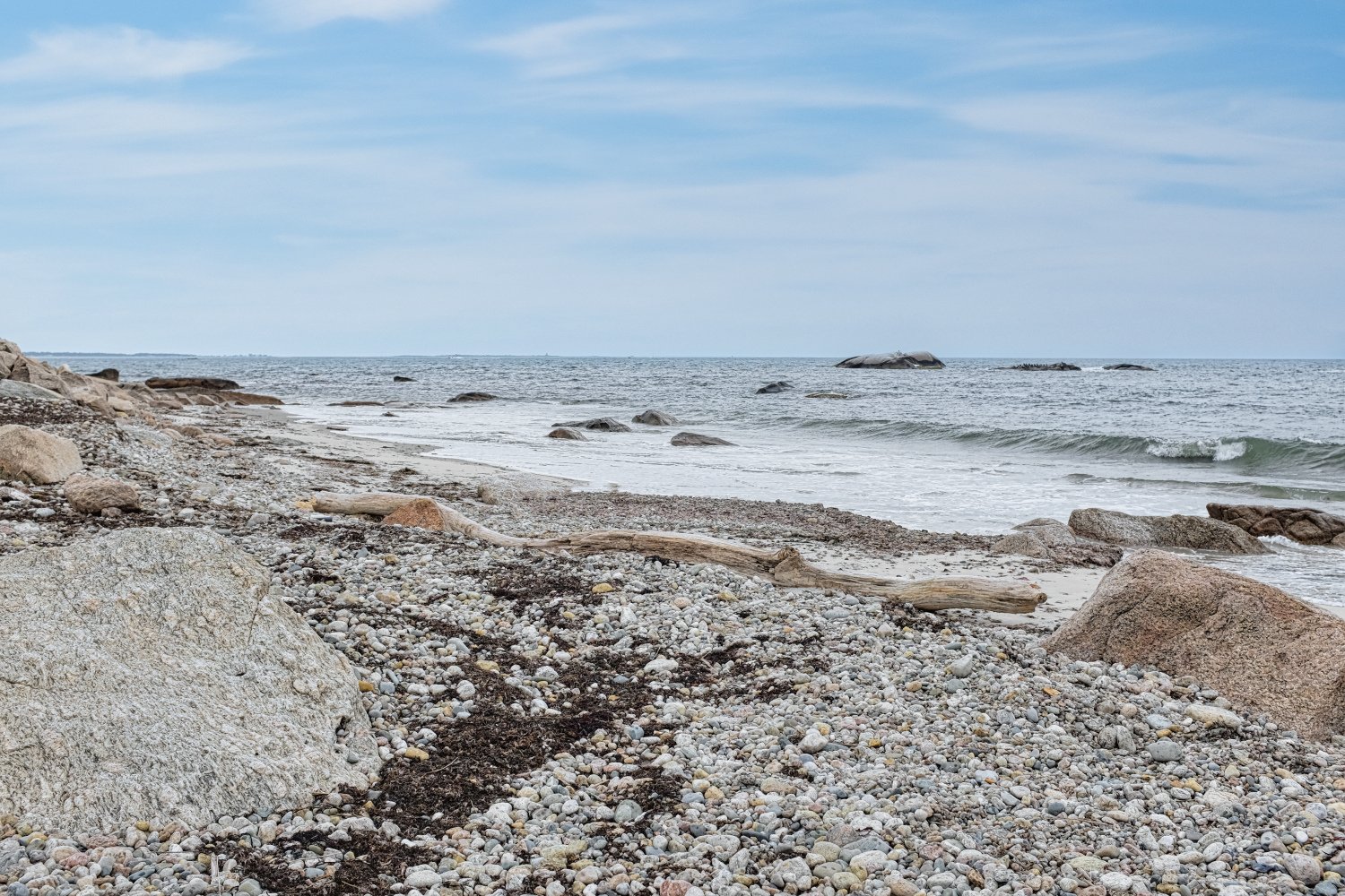 A rocky beach with small pebbles and driftwood, gentle waves, and an overcast sky over the ocean.