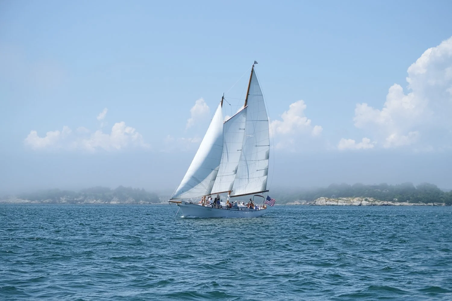 A sailboat with white sails on calm blue water near a distant shoreline with scattered trees and rocks, under a partly cloudy sky.