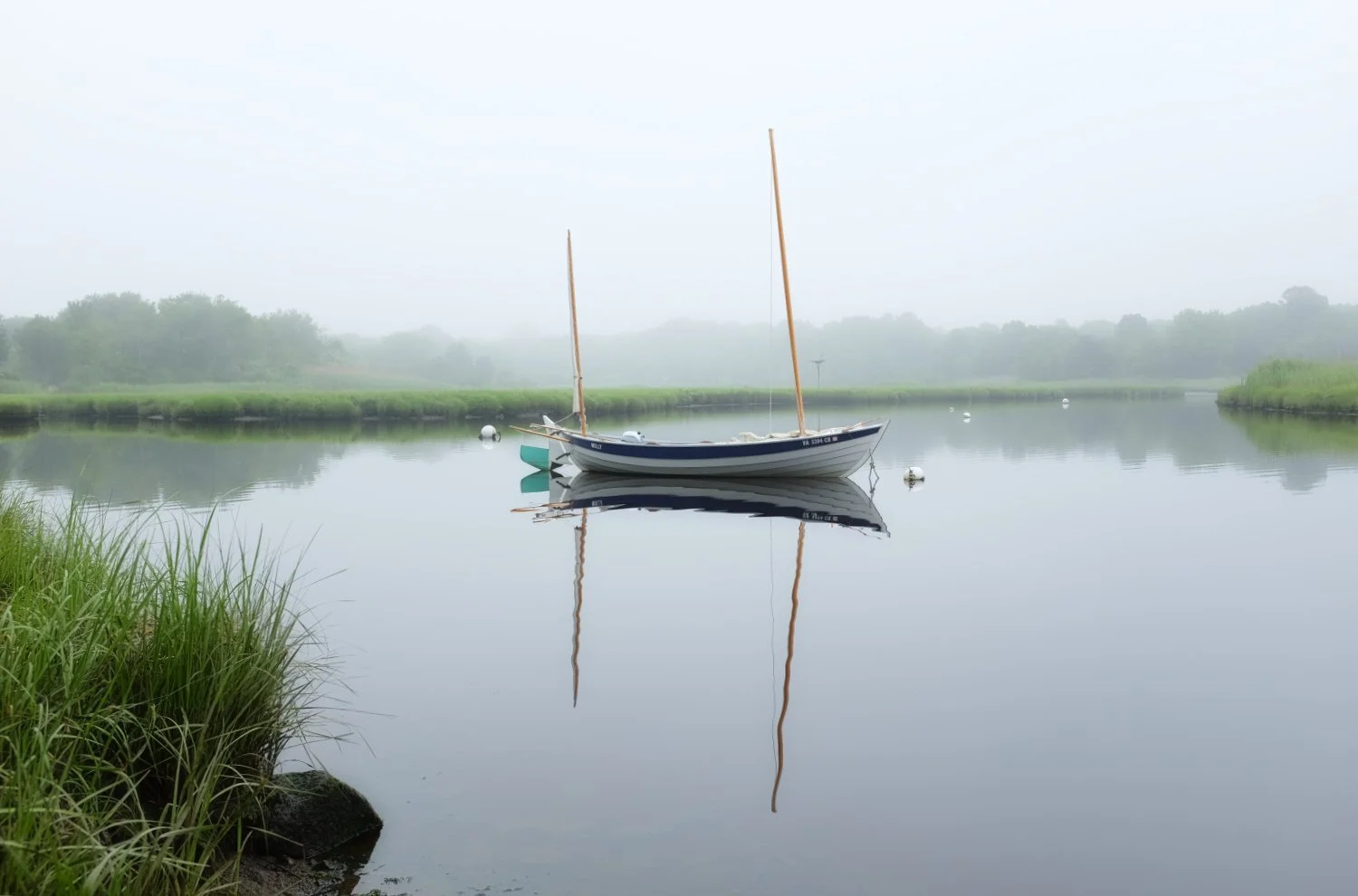 A sailboat floating on a calm, foggy river with green grassy banks and misty trees in the background.
