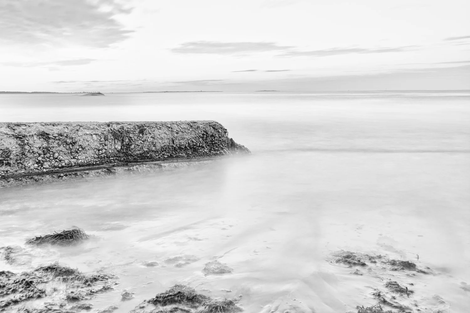 Black and white photograph of a rocky shoreline with a stone wall extending into calm water, with clouds and distant land visible in the background.