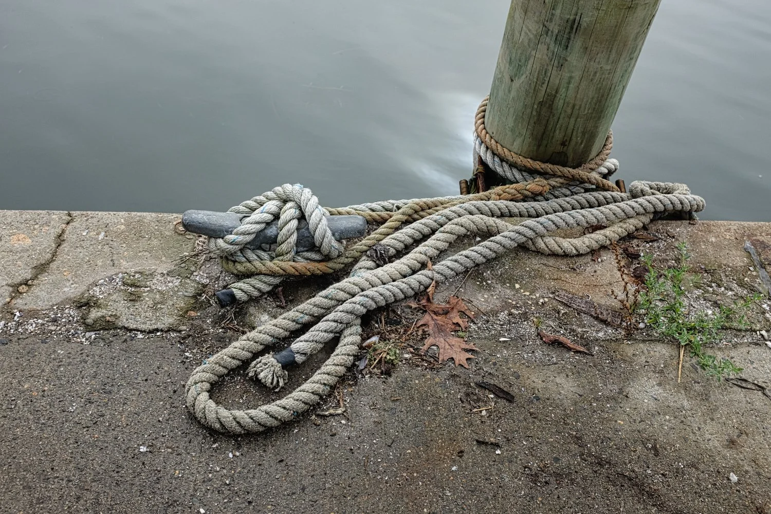Rope tied around a metal cleat on a concrete dock, with a boat mooring post nearby and water in the background.
