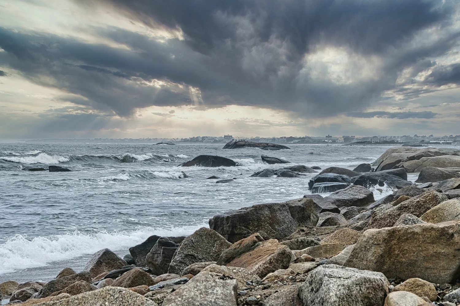 A rocky coastline with waves crashing against rocks under a cloudy, stormy sky, with distant houses visible across the water.
